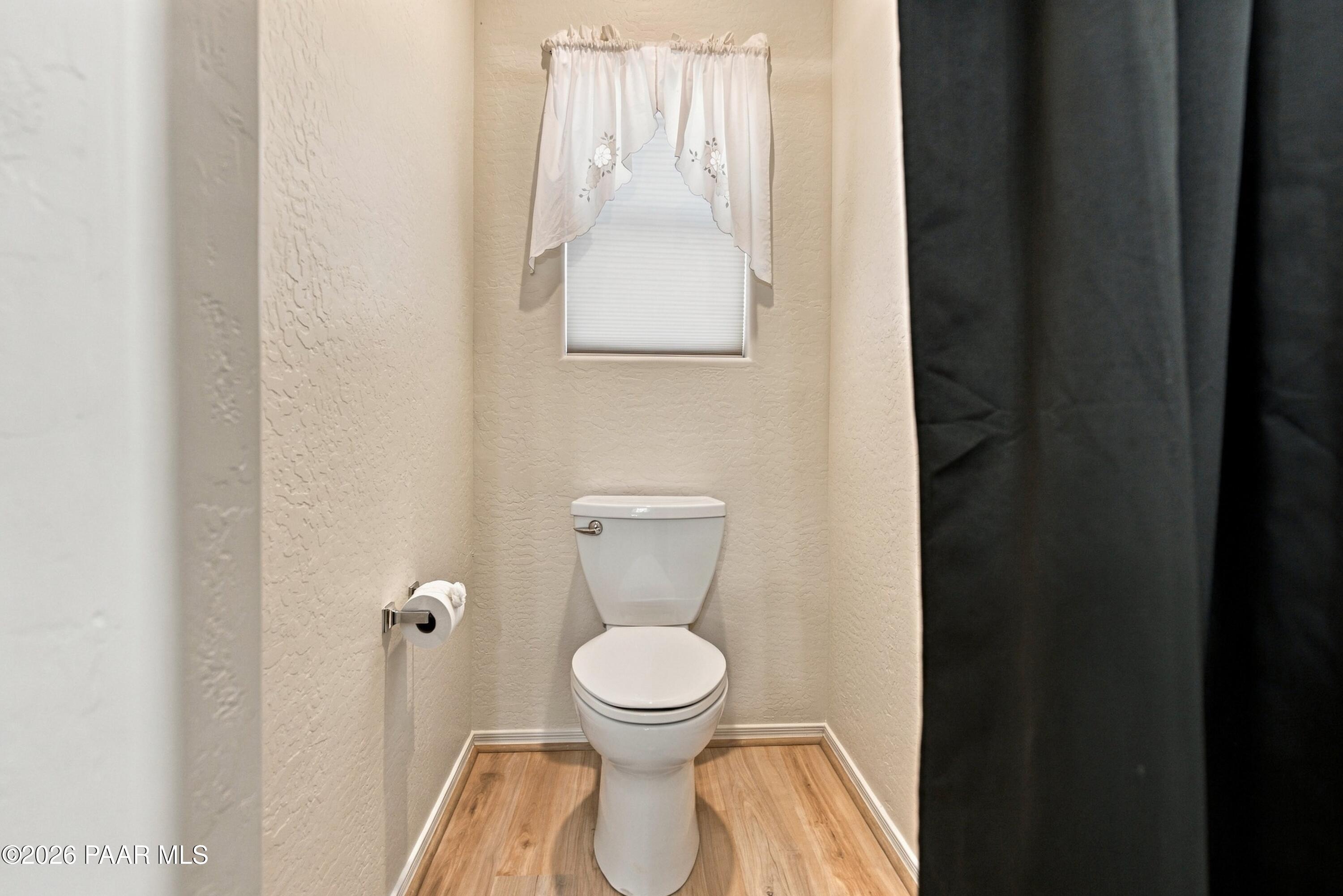 Toilet room with a white toilet beneath a curtained window and wood-look flooring.