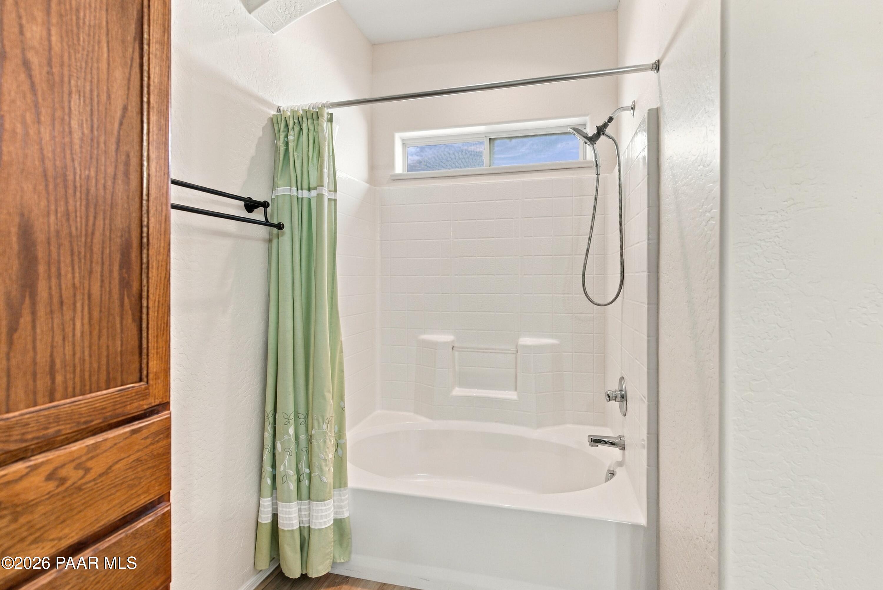 Bathroom with a white tub-shower, handheld showerhead, high window, and green patterned curtain.