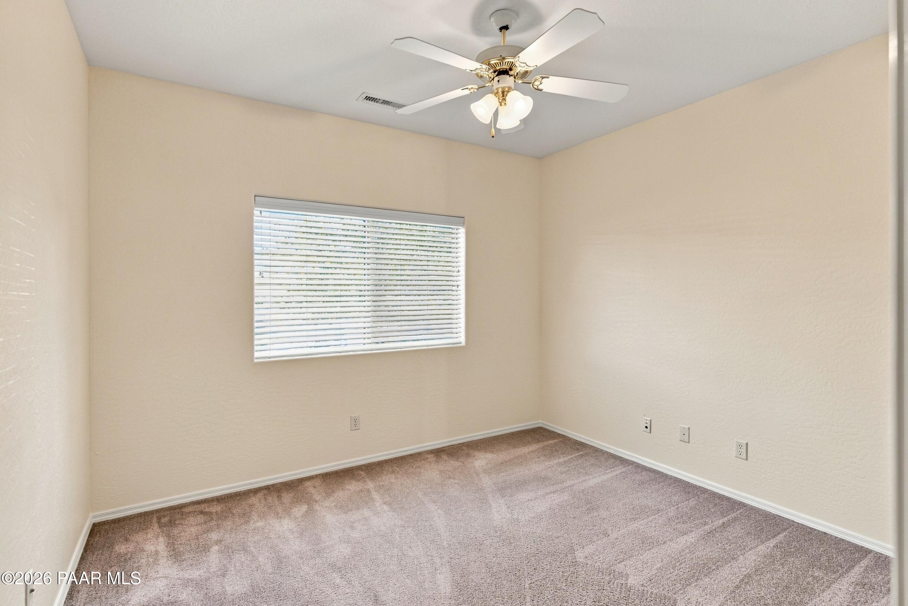 Empty bedroom with taupe carpet, a window with blinds, and a ceiling fan.