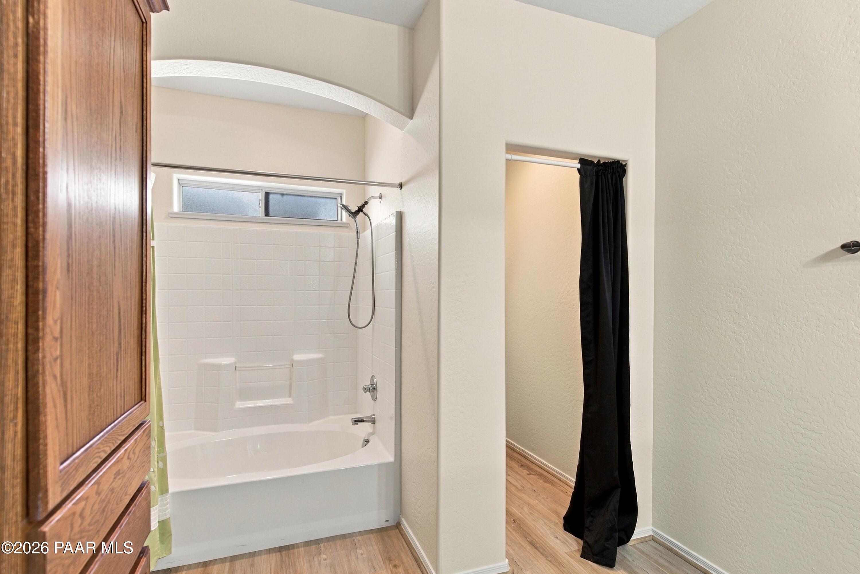 Bathroom with a white tub-shower enclosure, high window, wooden cabinet, and black doorway curtain.