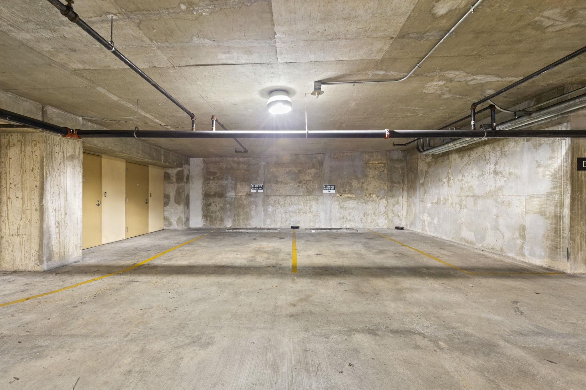 Indoor parking garage with two reserved resident spaces, yellow lines, and exposed concrete walls and pipes.