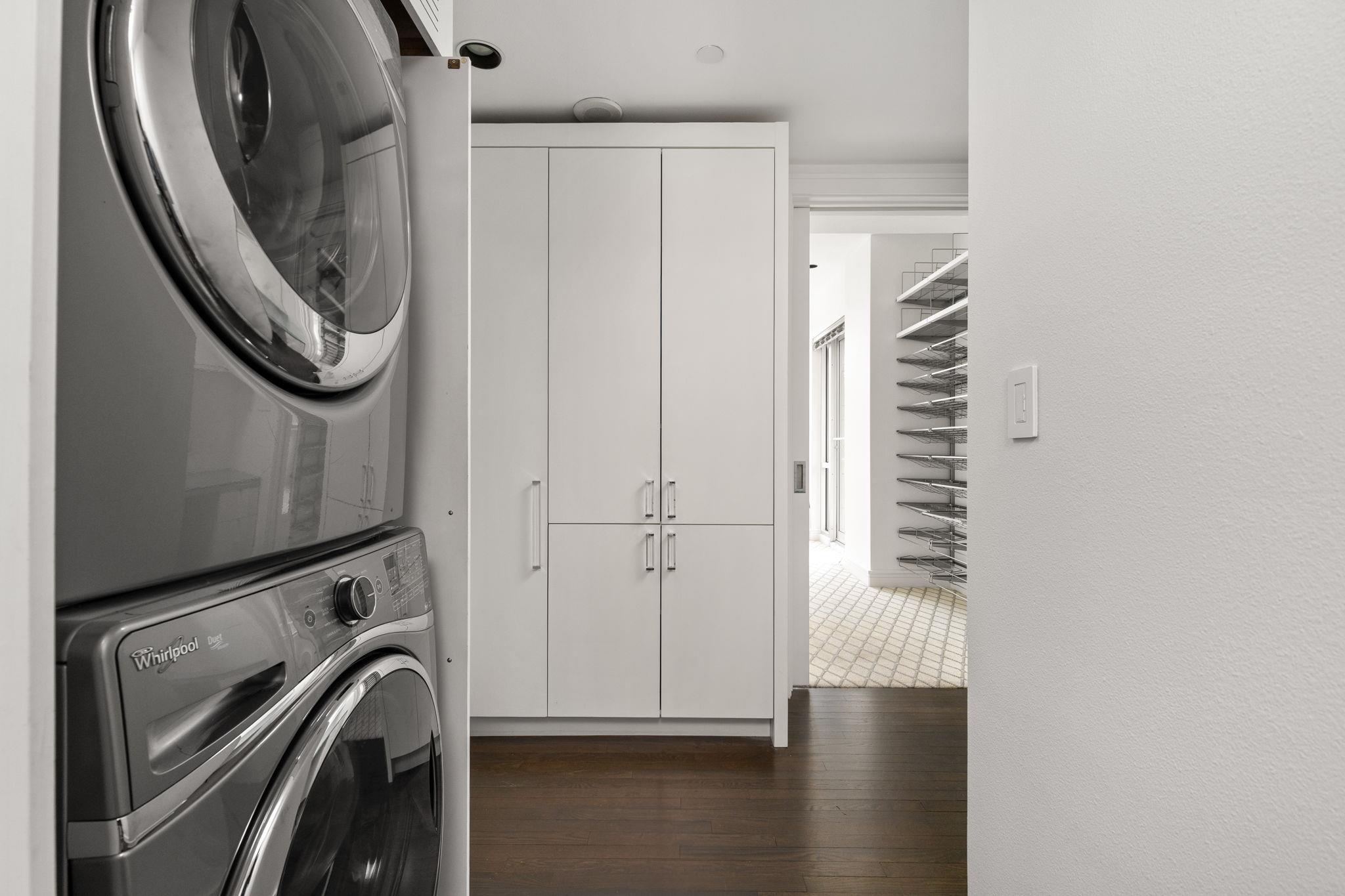 Laundry area with stacked front-load machines beside white cabinets, opening to a bright tiled space with wire shelving.
