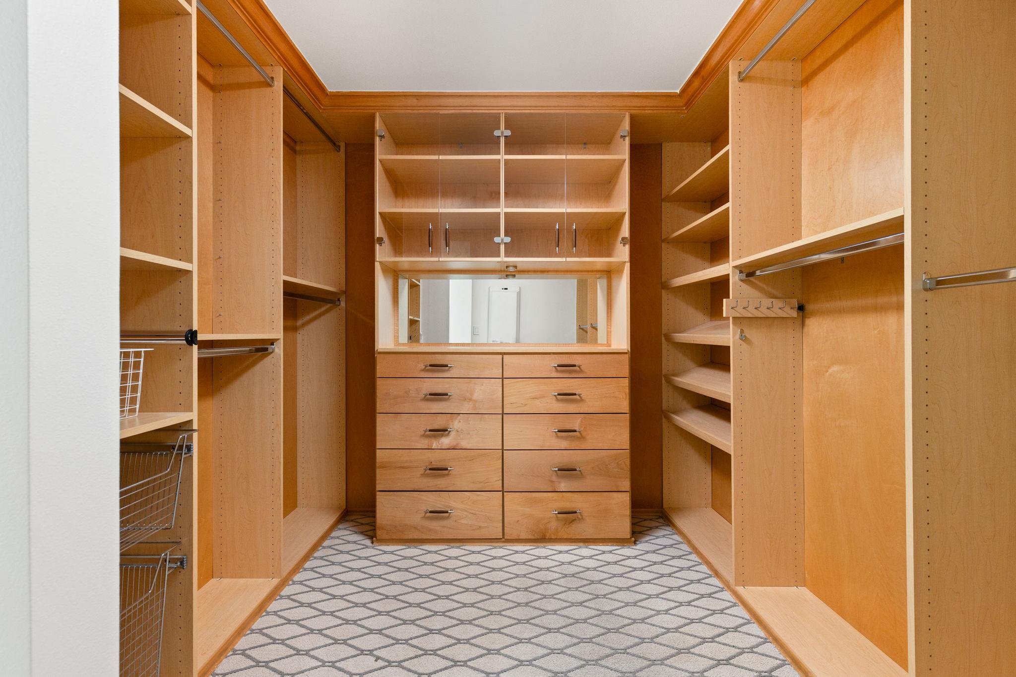 Walk-in closet with wood shelving, hanging rods, wire baskets, and central drawers beneath a mirror.