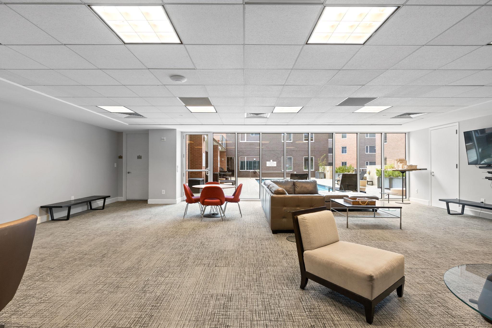 Furnished communal lounge with sofas and red dining chairs, overlooking a courtyard pool through large windows.