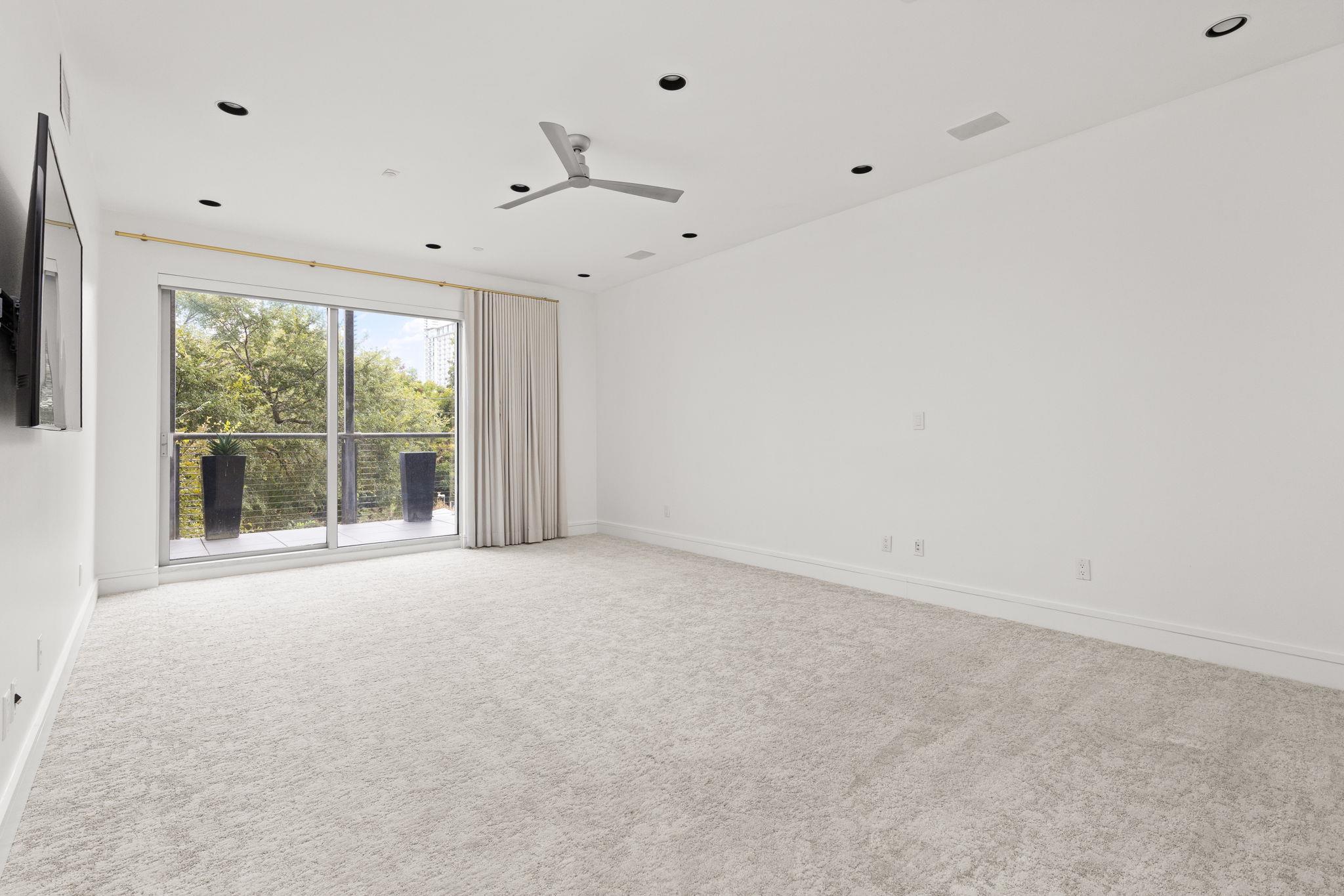 Empty living room with carpet, ceiling fan, and sliding doors opening to a tree-lined balcony.