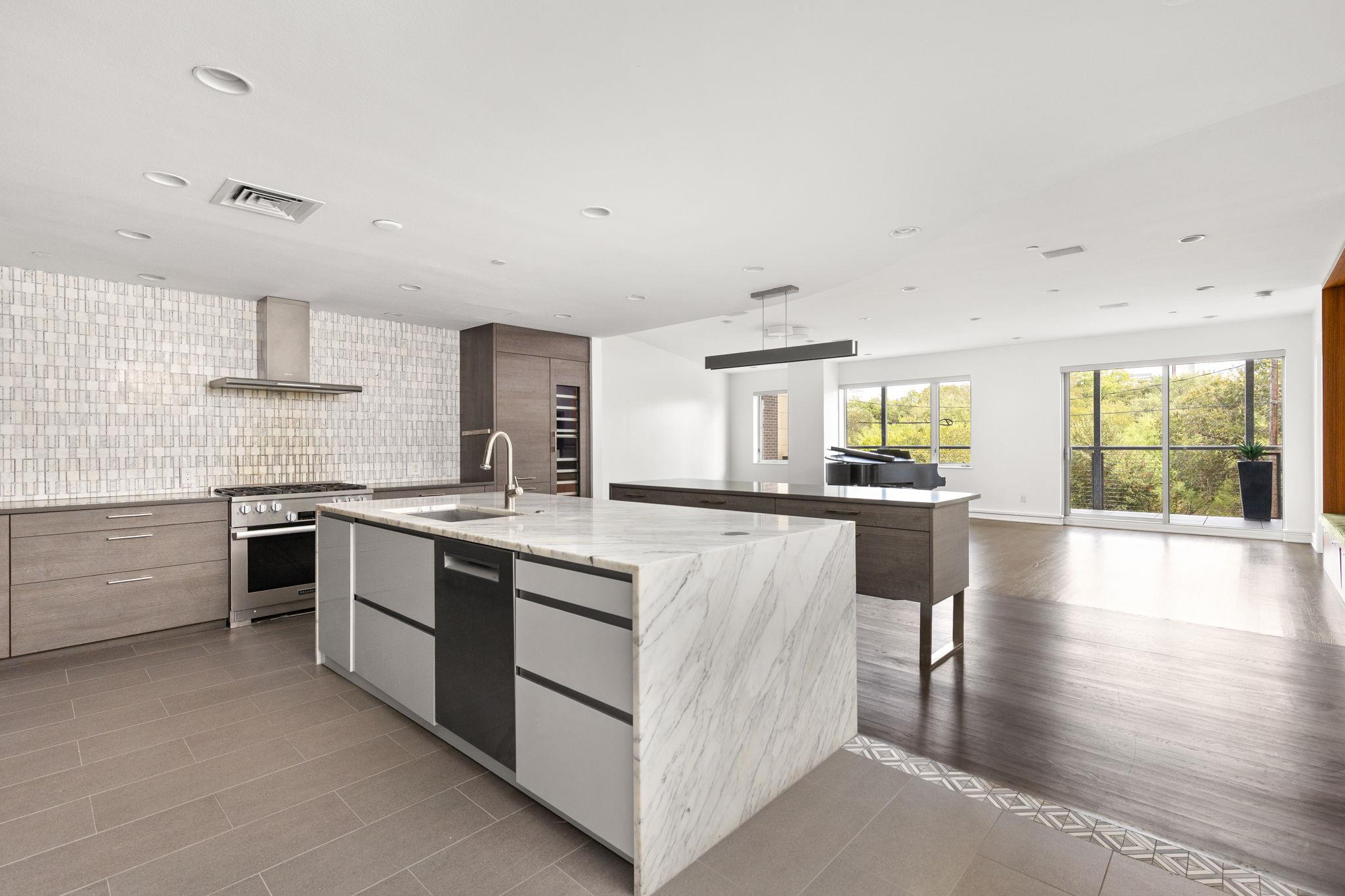 Modern kitchen with a large marble island and leafy views through floor-to-ceiling windows.