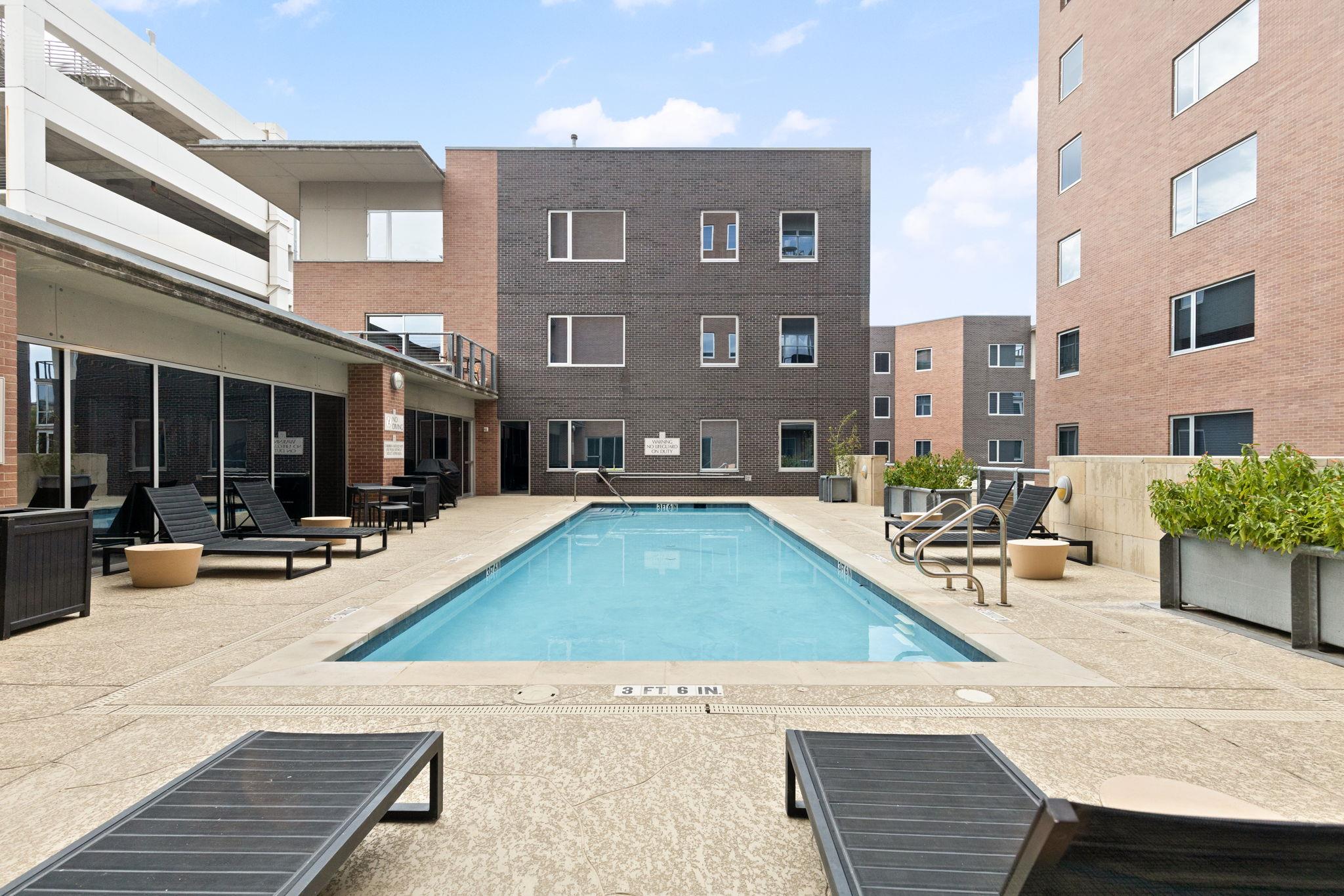 Outdoor swimming pool surrounded by brick buildings, lounge chairs, and planters.