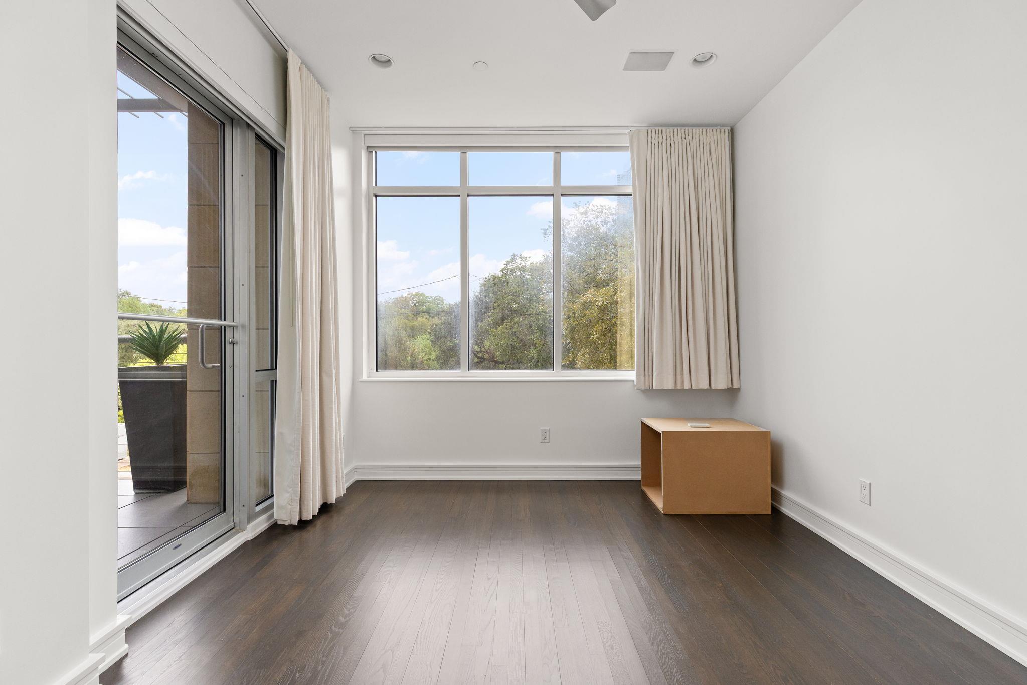Mostly empty room with dark wood floors, large curtained window, and sliding glass door to a balcony.