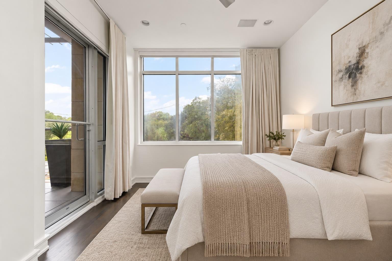 Staged bedroom with a large neutral bed, broad windows, and sliding glass doors to a balcony.