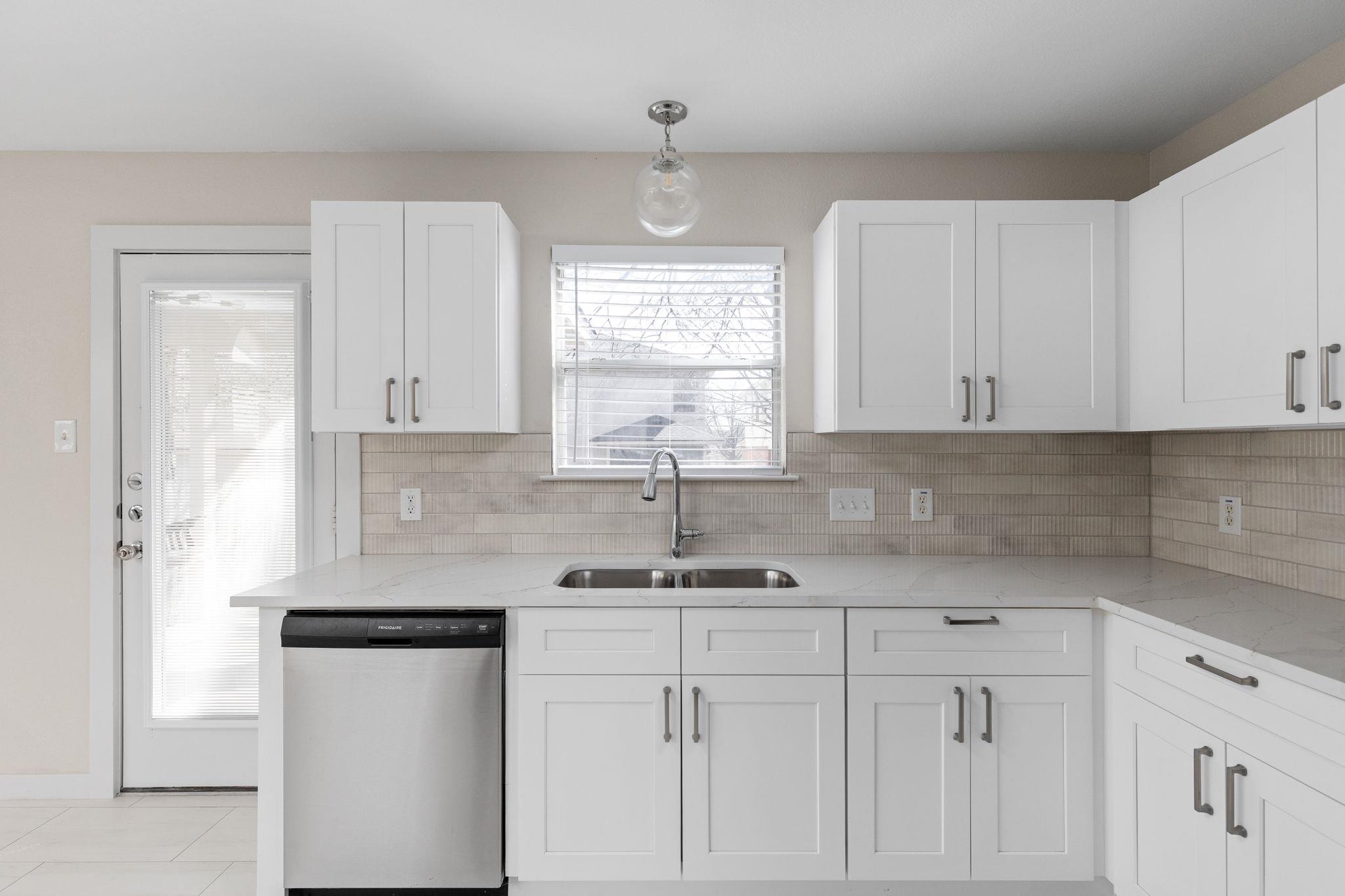 Kitchen with white cabinets, veined counters, a tiled backsplash, and a double sink beneath the window.