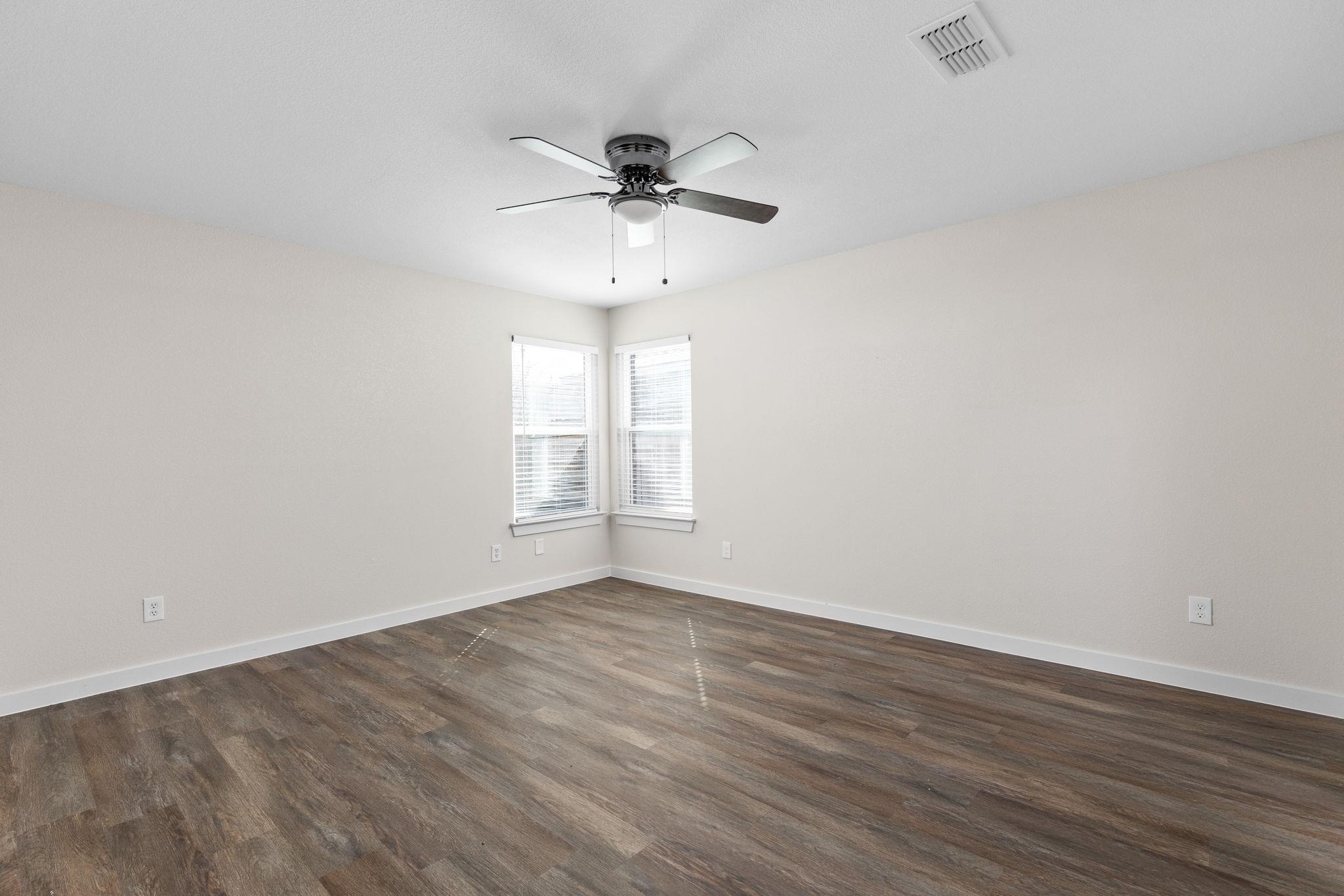 Empty bedroom with dark plank flooring, corner windows with blinds, and a ceiling fan.