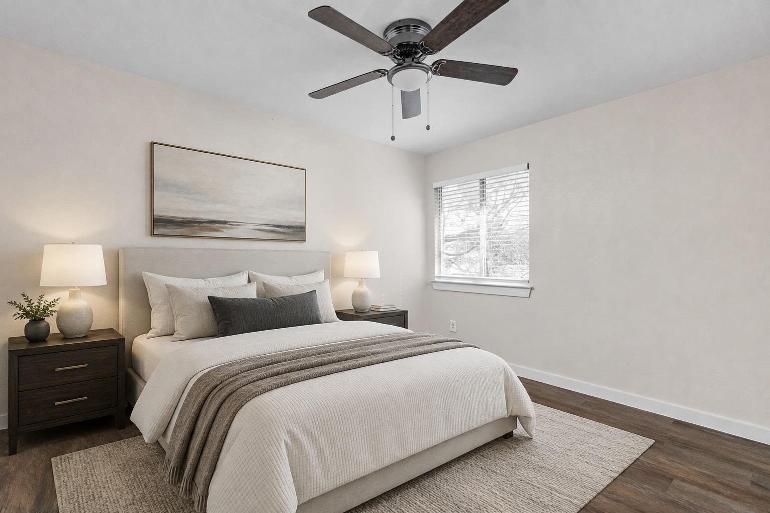 Furnished bedroom with a large upholstered bed, layered neutral bedding, dark wood nightstands, and a ceiling fan.