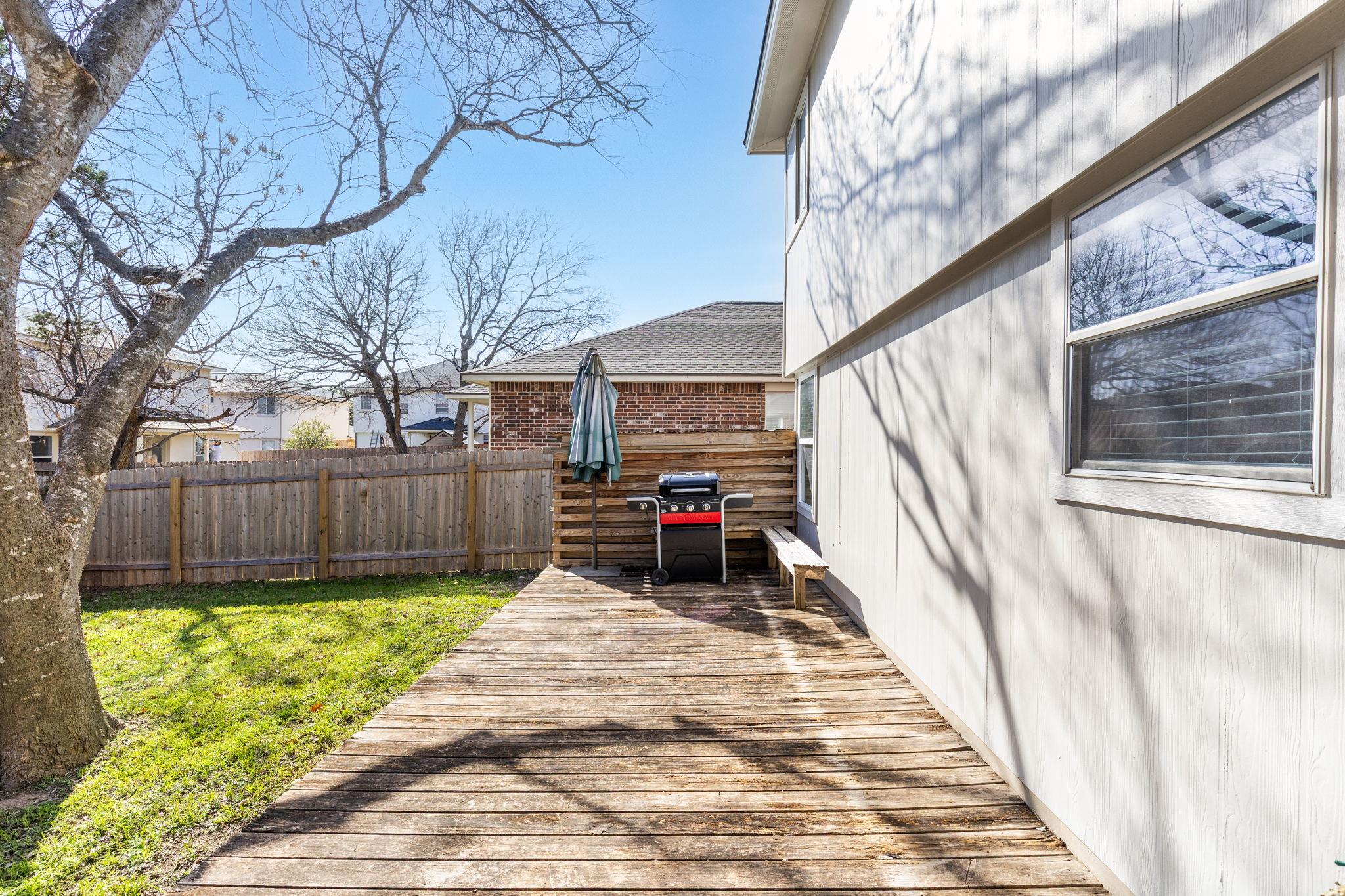 Backyard deck beside a light-sided house, with a grill, closed umbrella, fenced lawn, and large leafless tree.