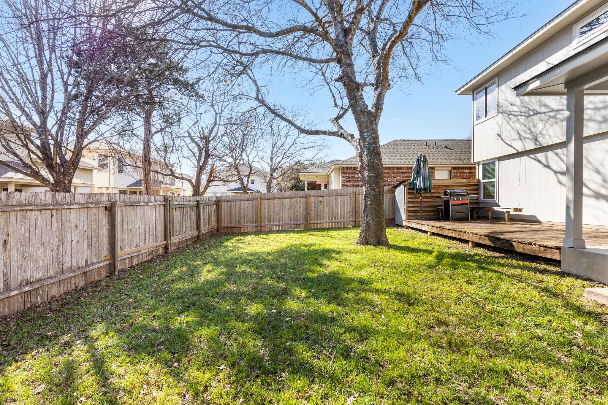 Fenced backyard with a large tree, green lawn, and raised deck with a grill.