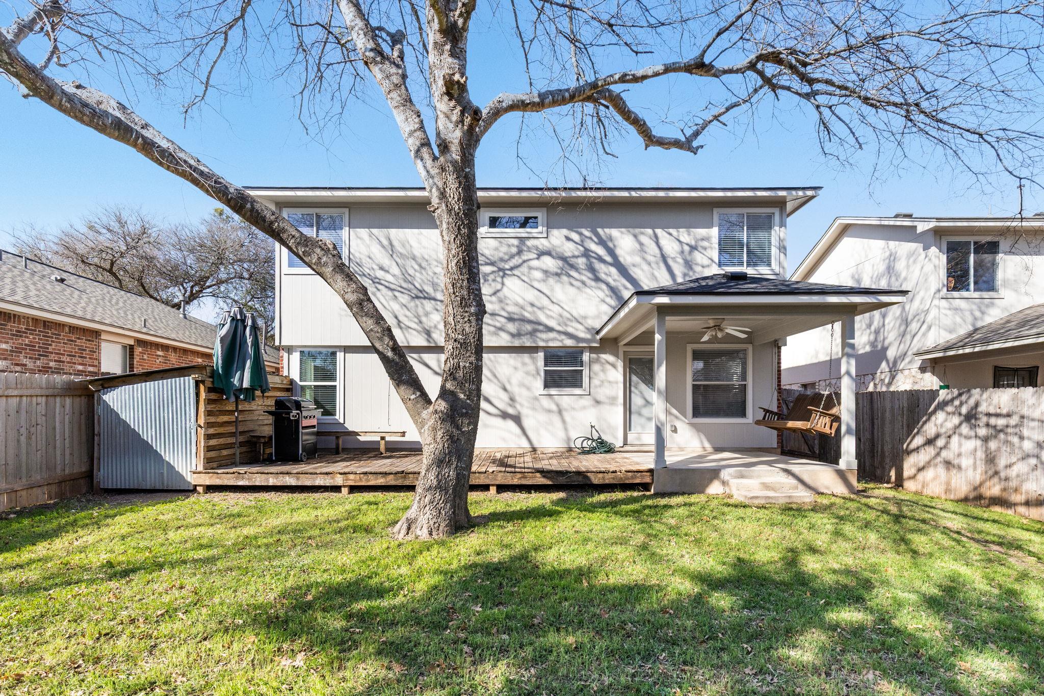 Rear of a two-story house with a covered patio, wooden deck, and large tree in the grassy backyard.