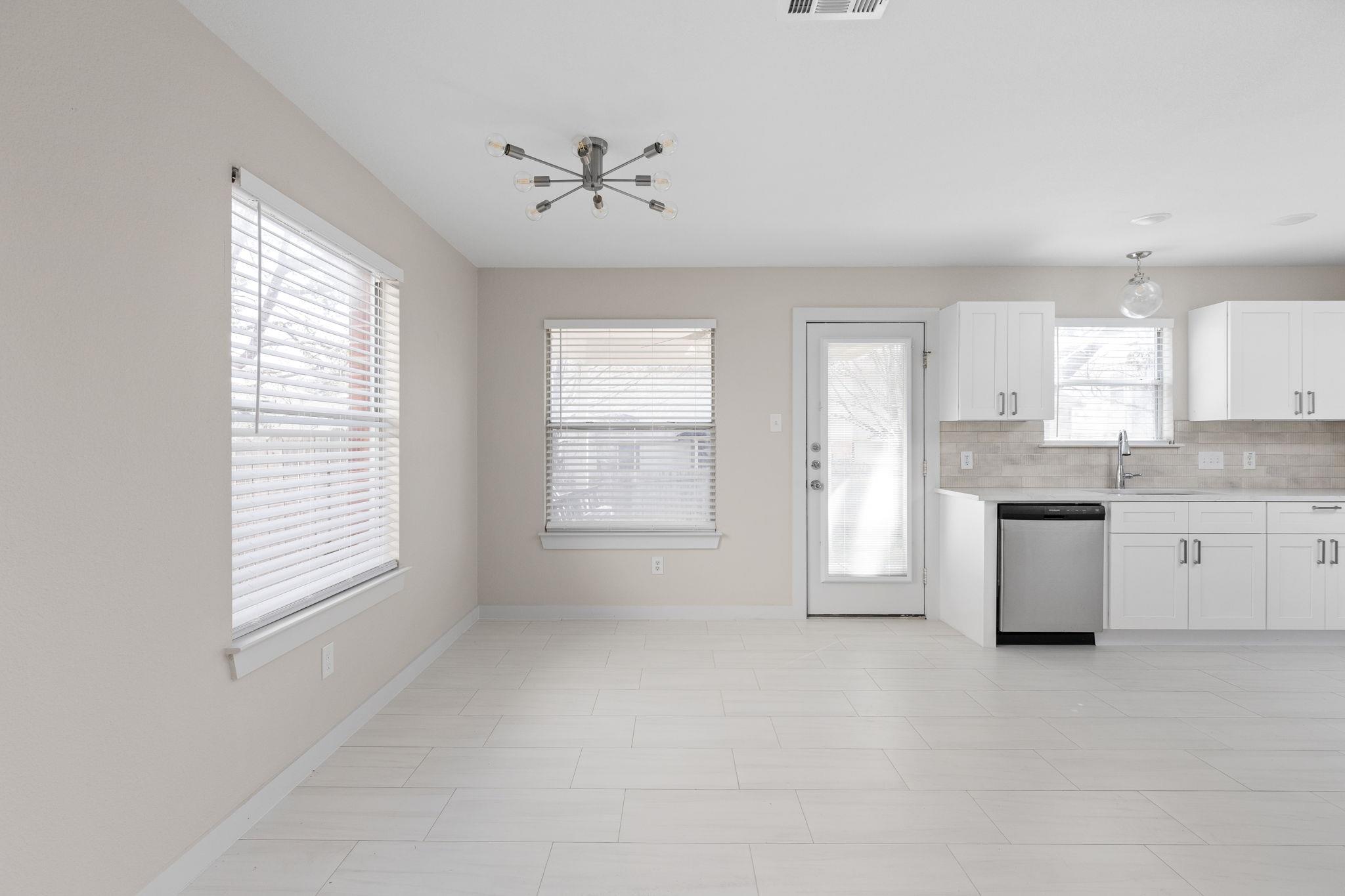 Bright vacant kitchen with white cabinets, stainless-steel dishwasher, tiled floor, and glass-paneled exterior door.