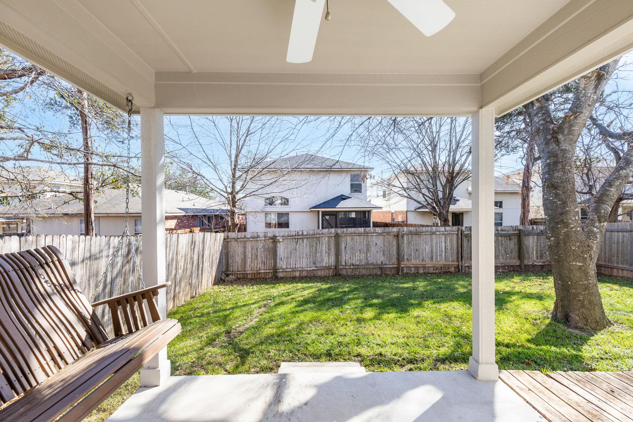 Covered backyard patio with a wooden swing overlooking a grassy, fenced yard and mature tree.