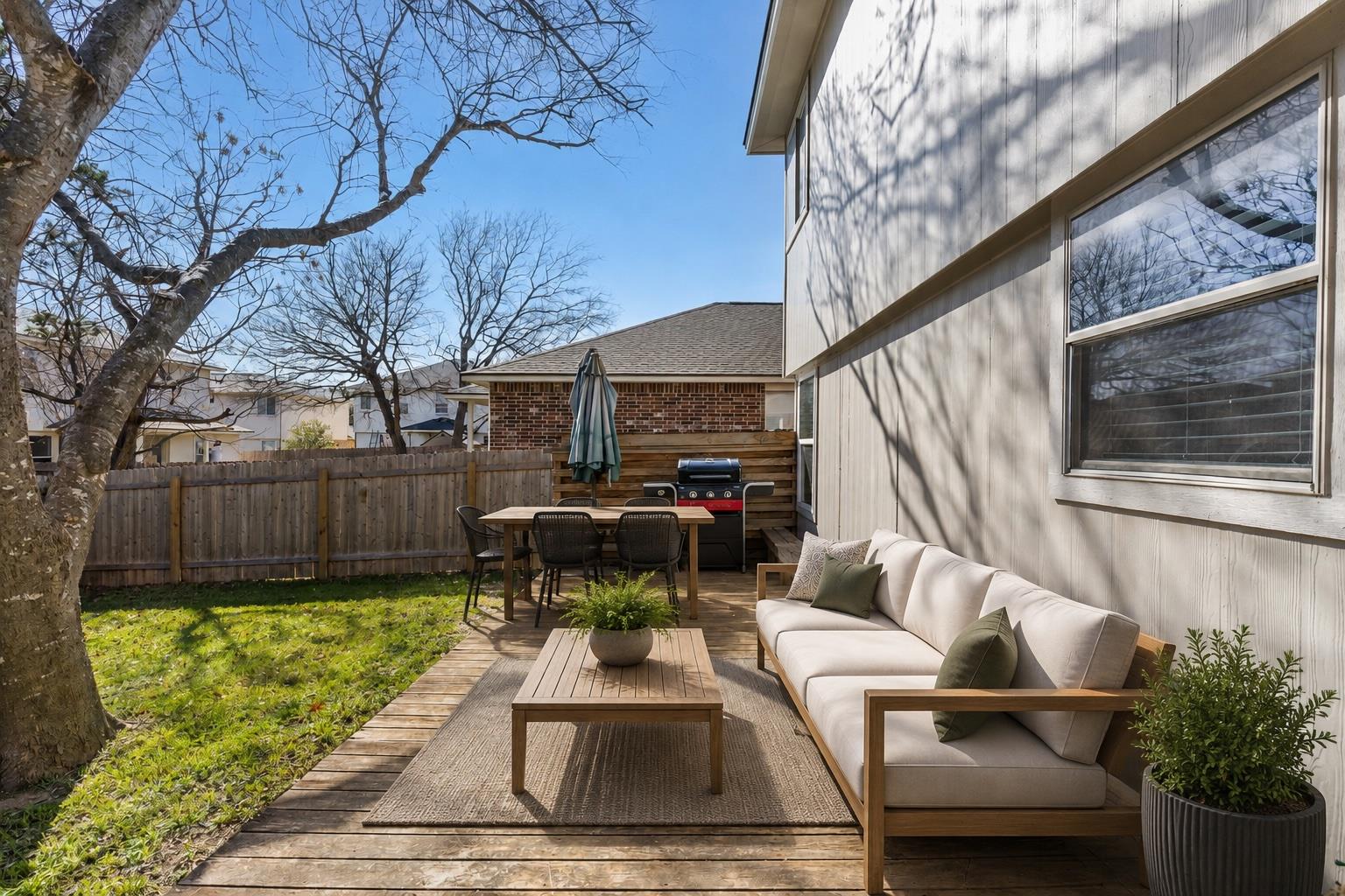 Backyard patio with a wood deck, outdoor sofa, dining set, and grill beside a fenced lawn.