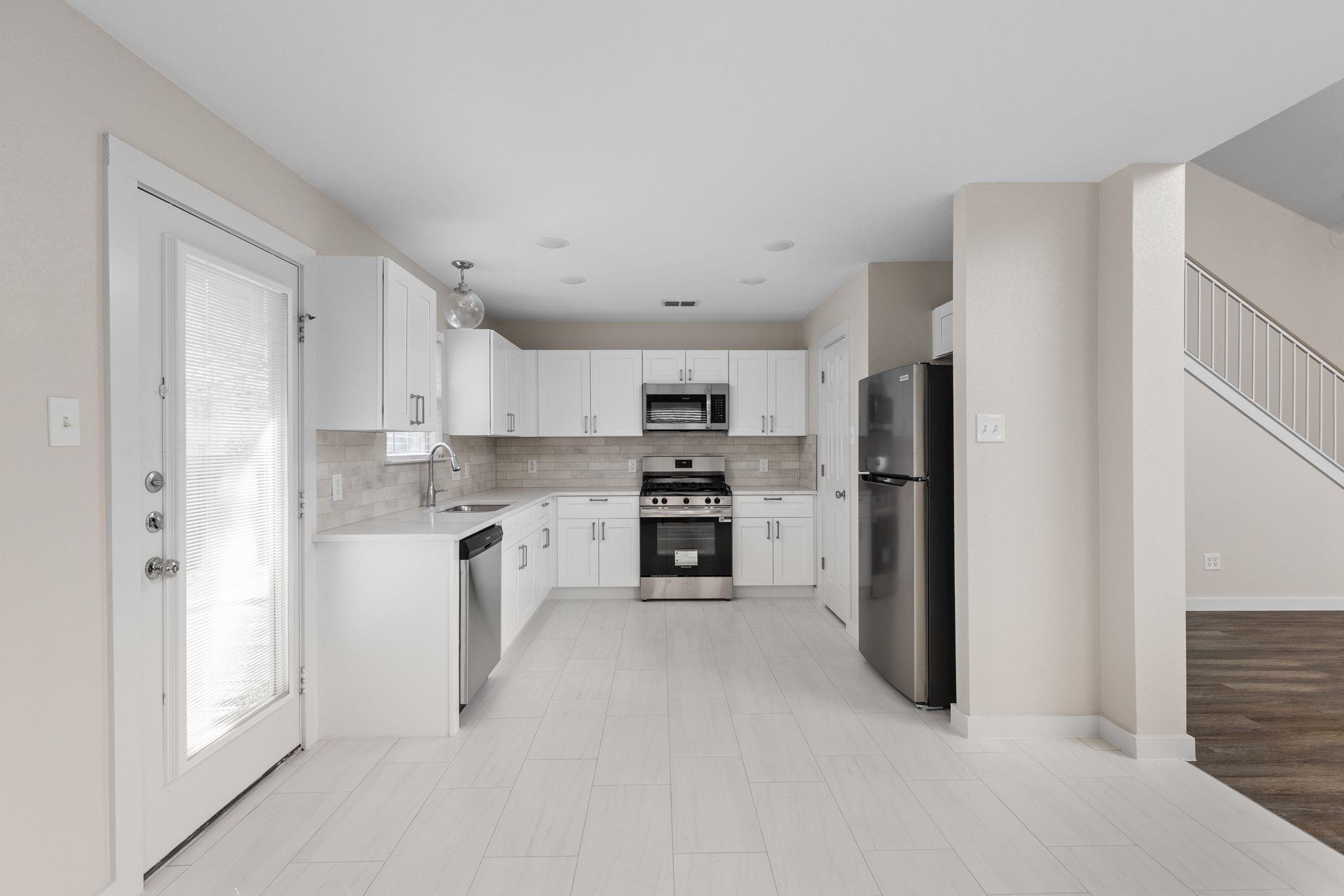 Bright white kitchen with stainless steel appliances, tile floors, and an adjacent staircase.