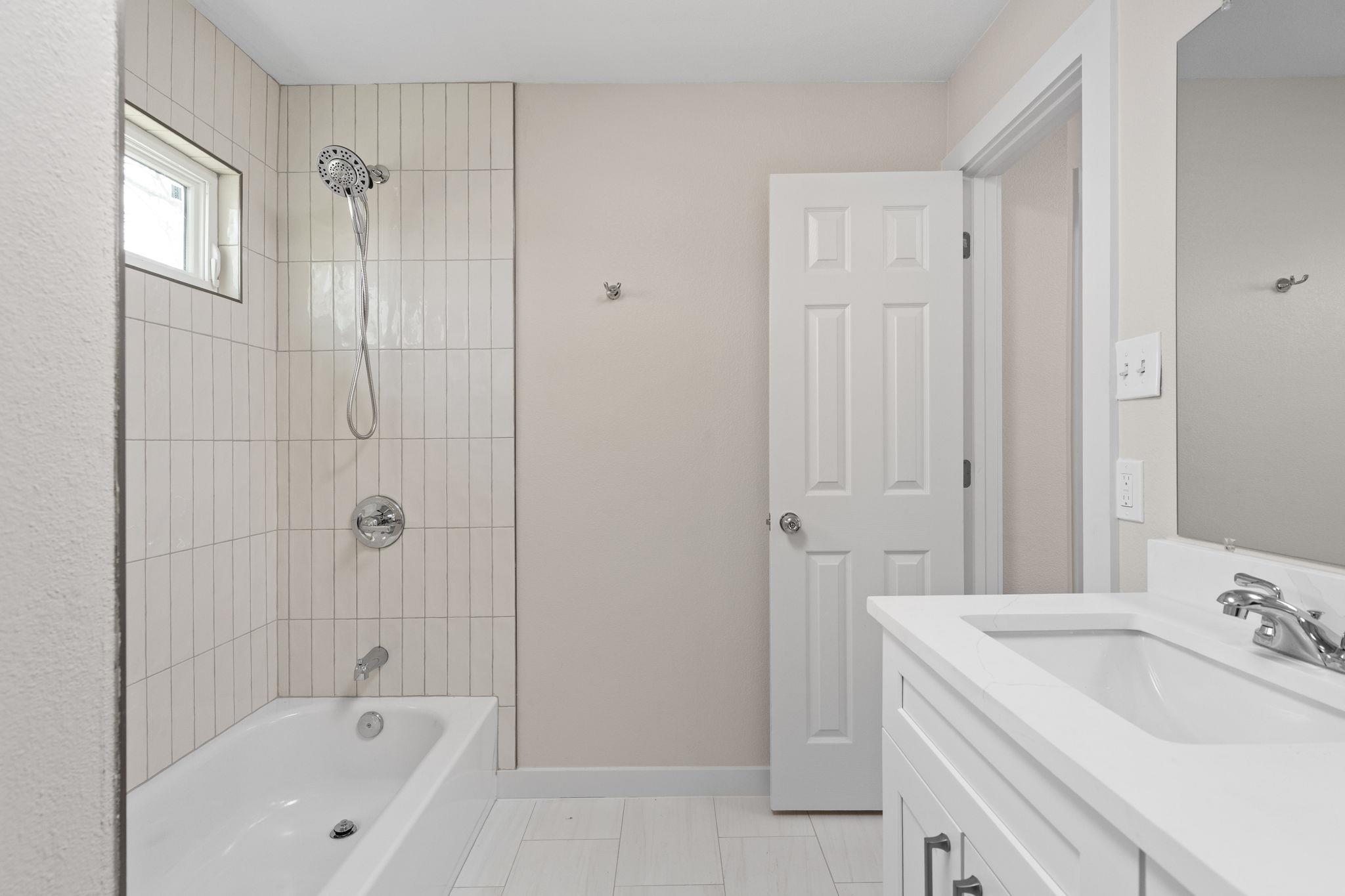 Bathroom with a white tub, tiled shower, small window, and white vanity.