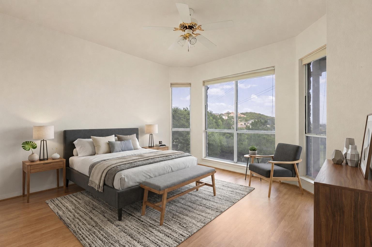 Staged bedroom with a gray bed, wooden furniture, and large windows overlooking leafy hills.