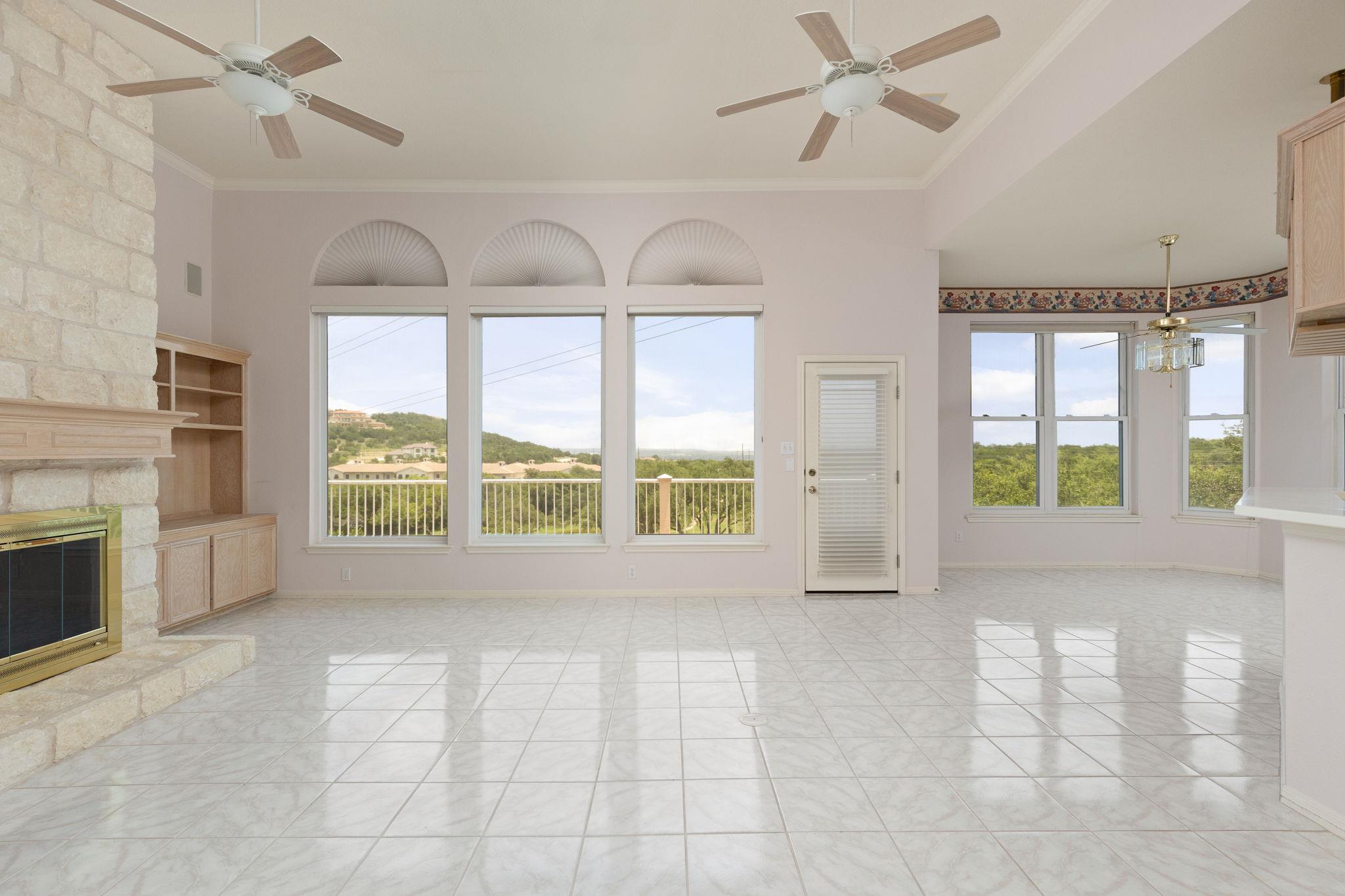 Vacant living room with a stone fireplace, tiled floors, and large windows overlooking green hills.