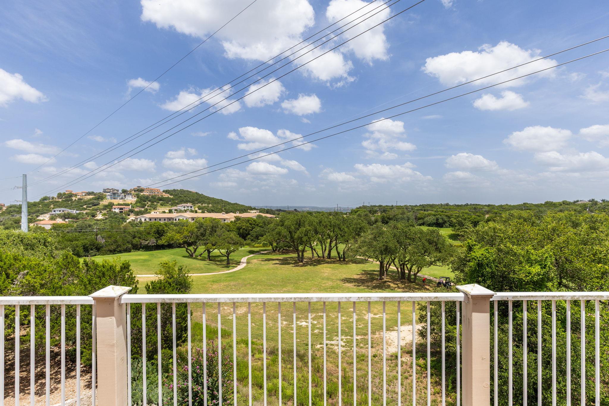 Golf course view from a balcony, with a winding path, trees, and homes on a distant hill.