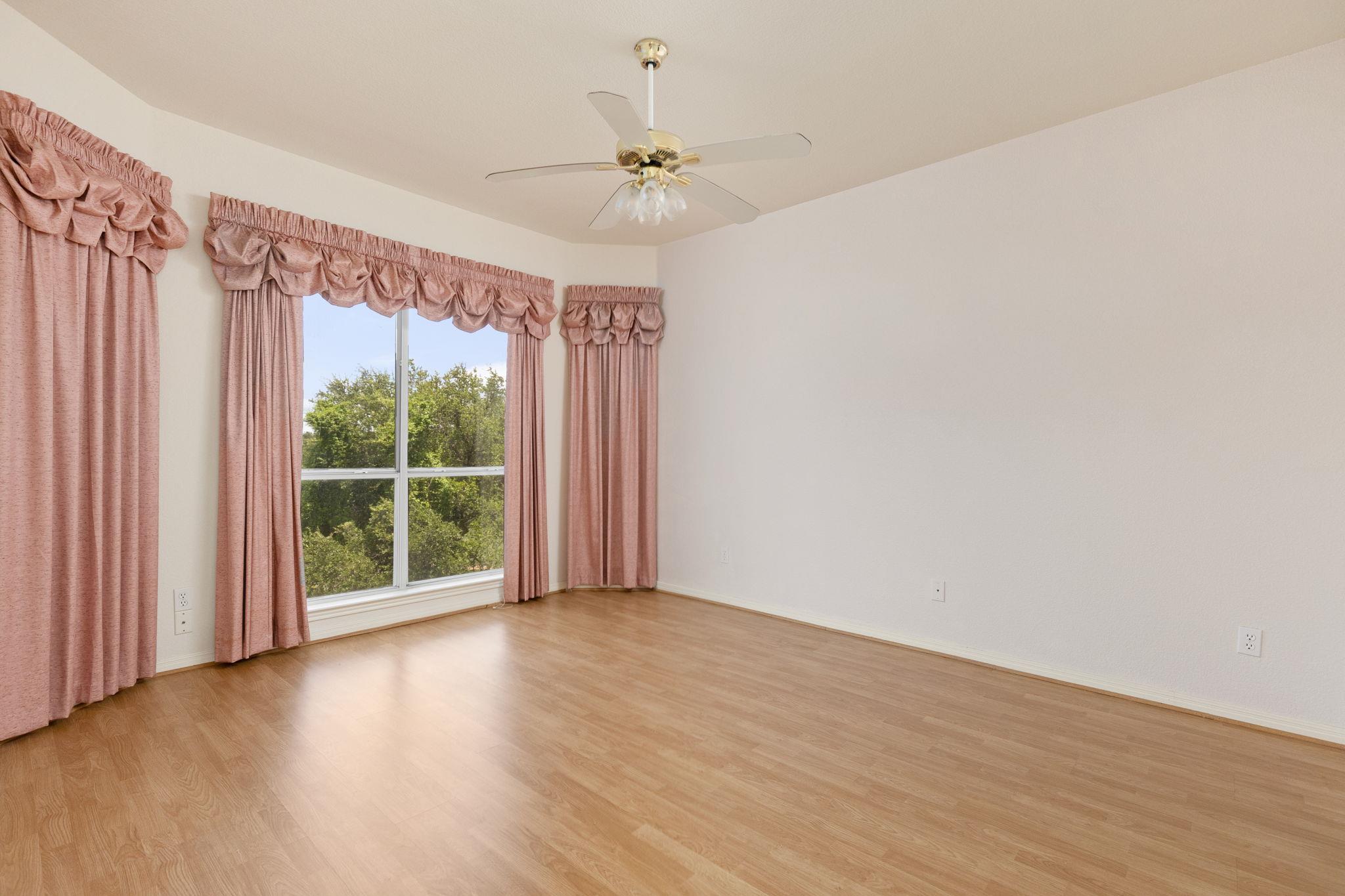 Empty room with wood flooring, pink curtains, and a large window overlooking trees.