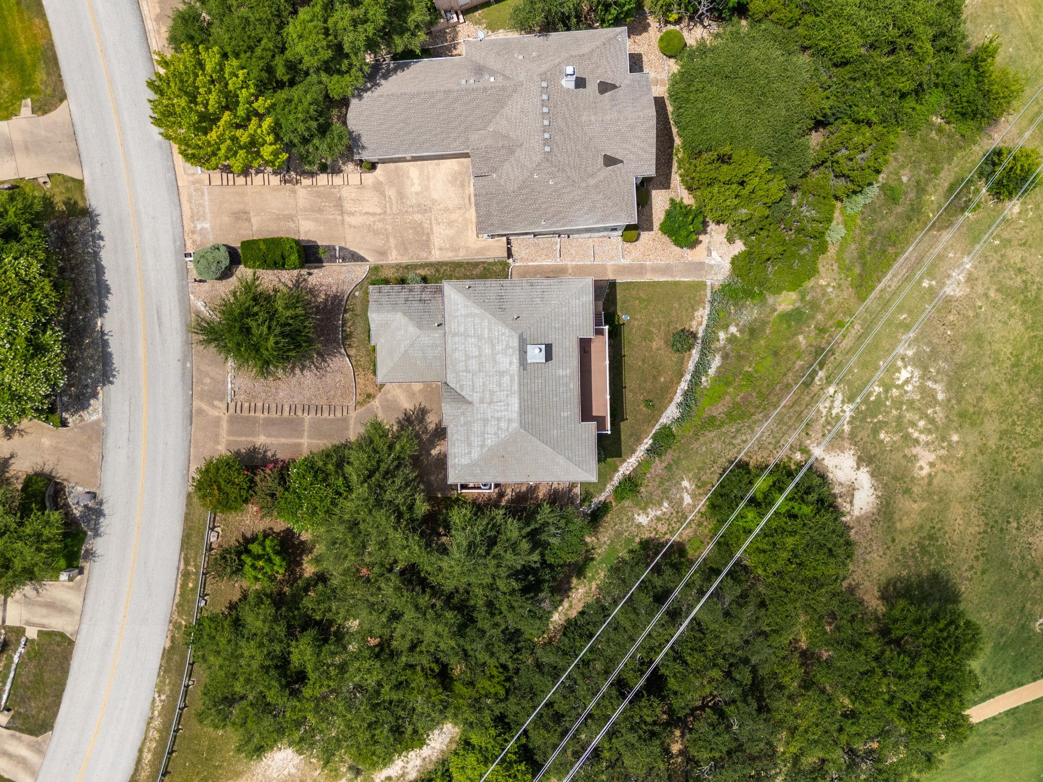 Aerial view of a gray-roofed house beside a road, surrounded by mature trees and open green grounds.