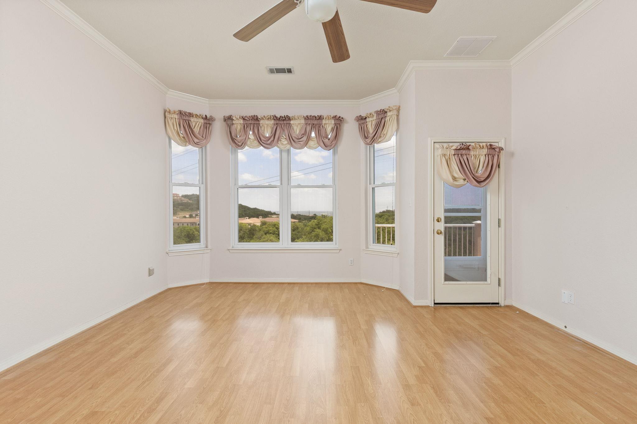 Vacant room with light wood flooring, bay windows overlooking green hills, and a glass-paneled balcony door.