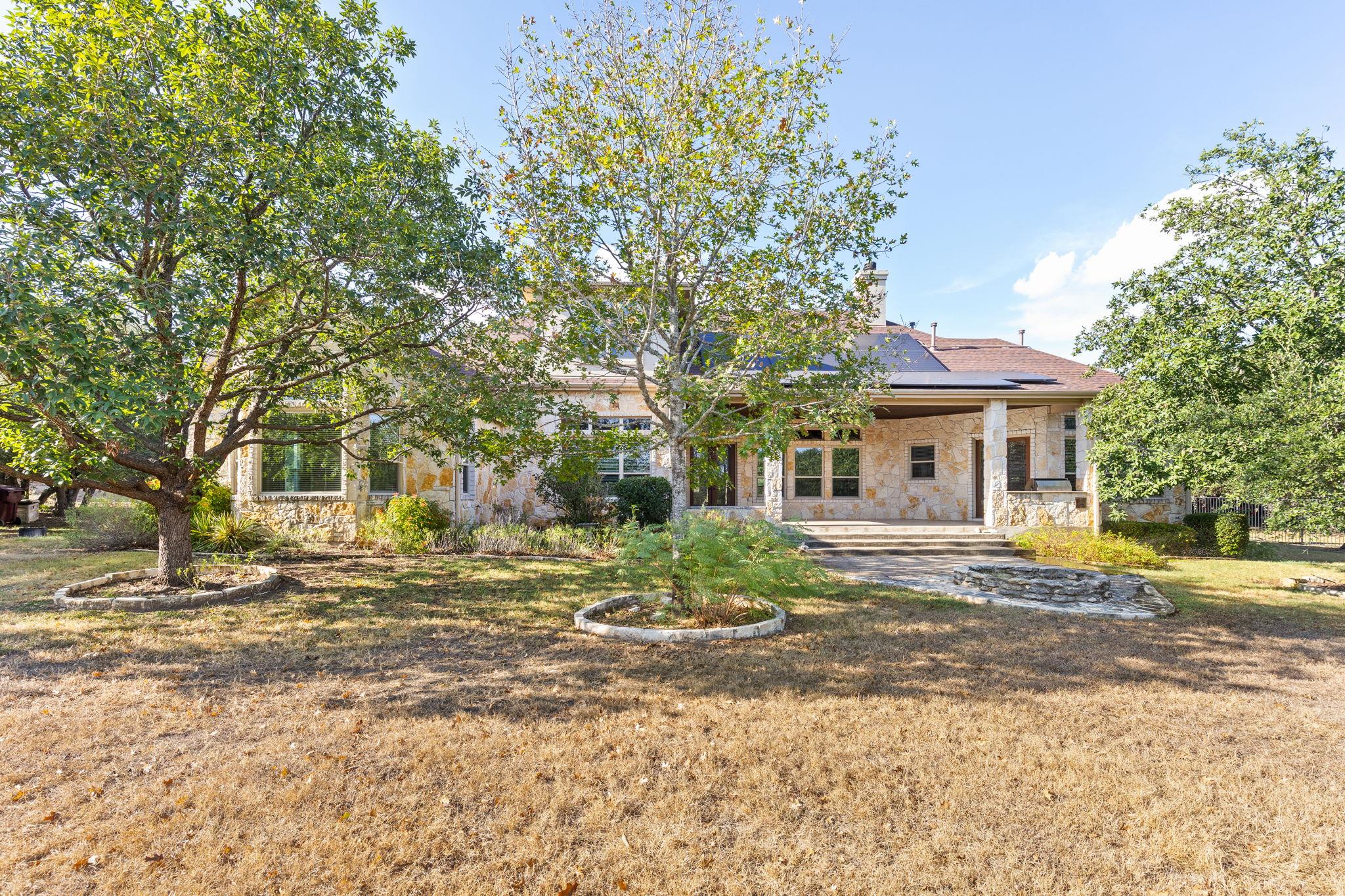 Stone home with a covered patio and solar panels, framed by mature trees and a dry lawn.