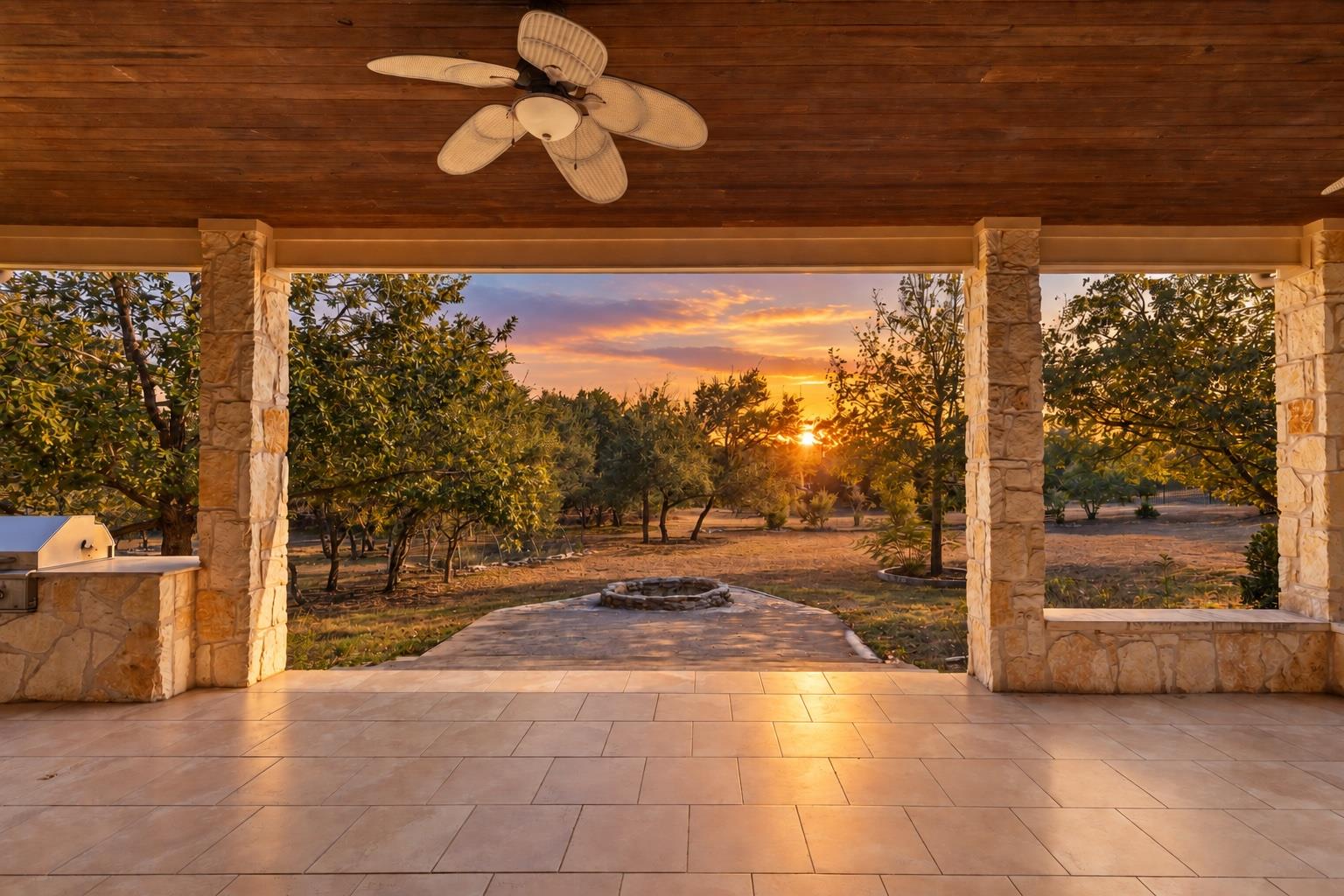 Covered stone patio overlooking a tree-filled yard at sunset, with a ceiling fan and built-in grill.