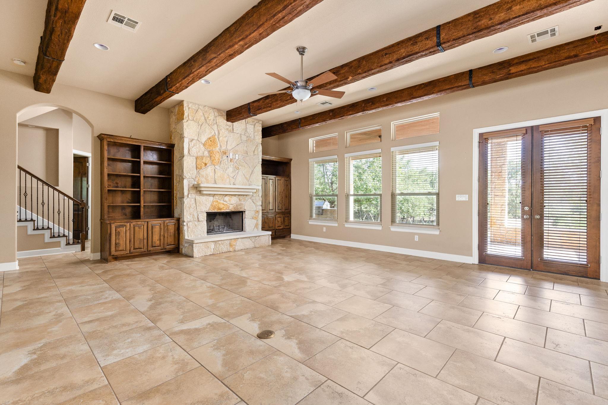 Empty living room with a stone fireplace, exposed wood beams, built-in shelving, and tile floors.