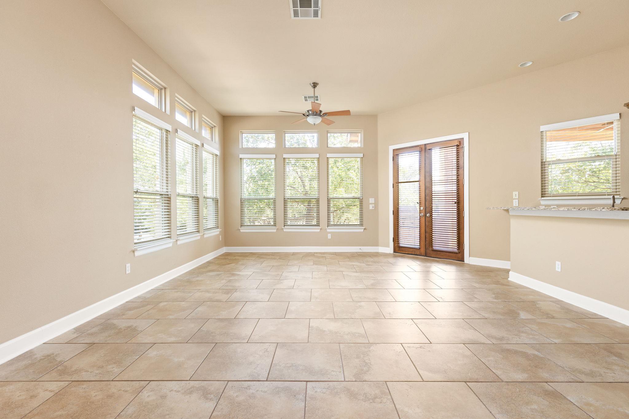 Empty dining area with large windows, tan tile flooring, and wooden double doors with blinds.