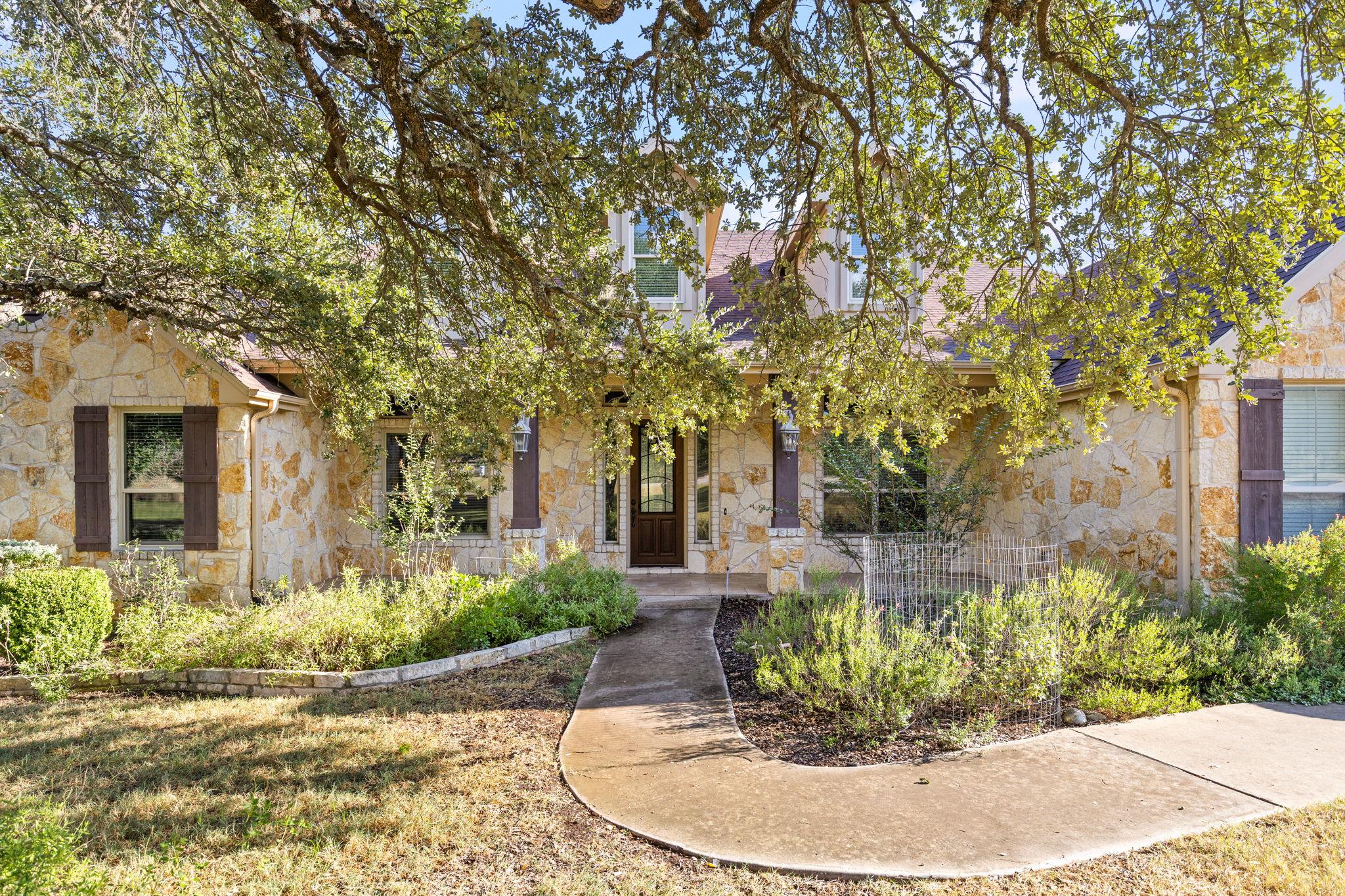Stone house entrance beneath a spreading tree, with a curved walkway and landscaped garden beds.