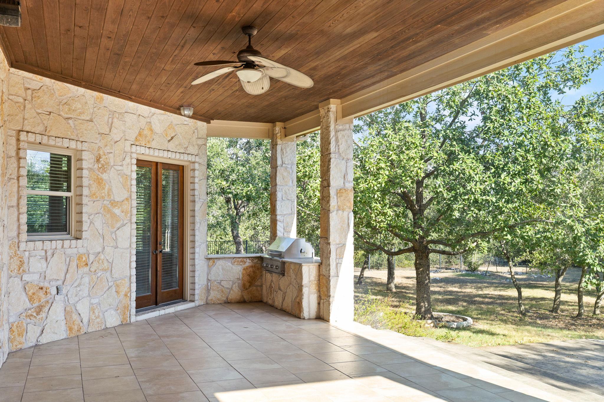 Covered stone patio with a built-in grill and mature oak trees beyond.