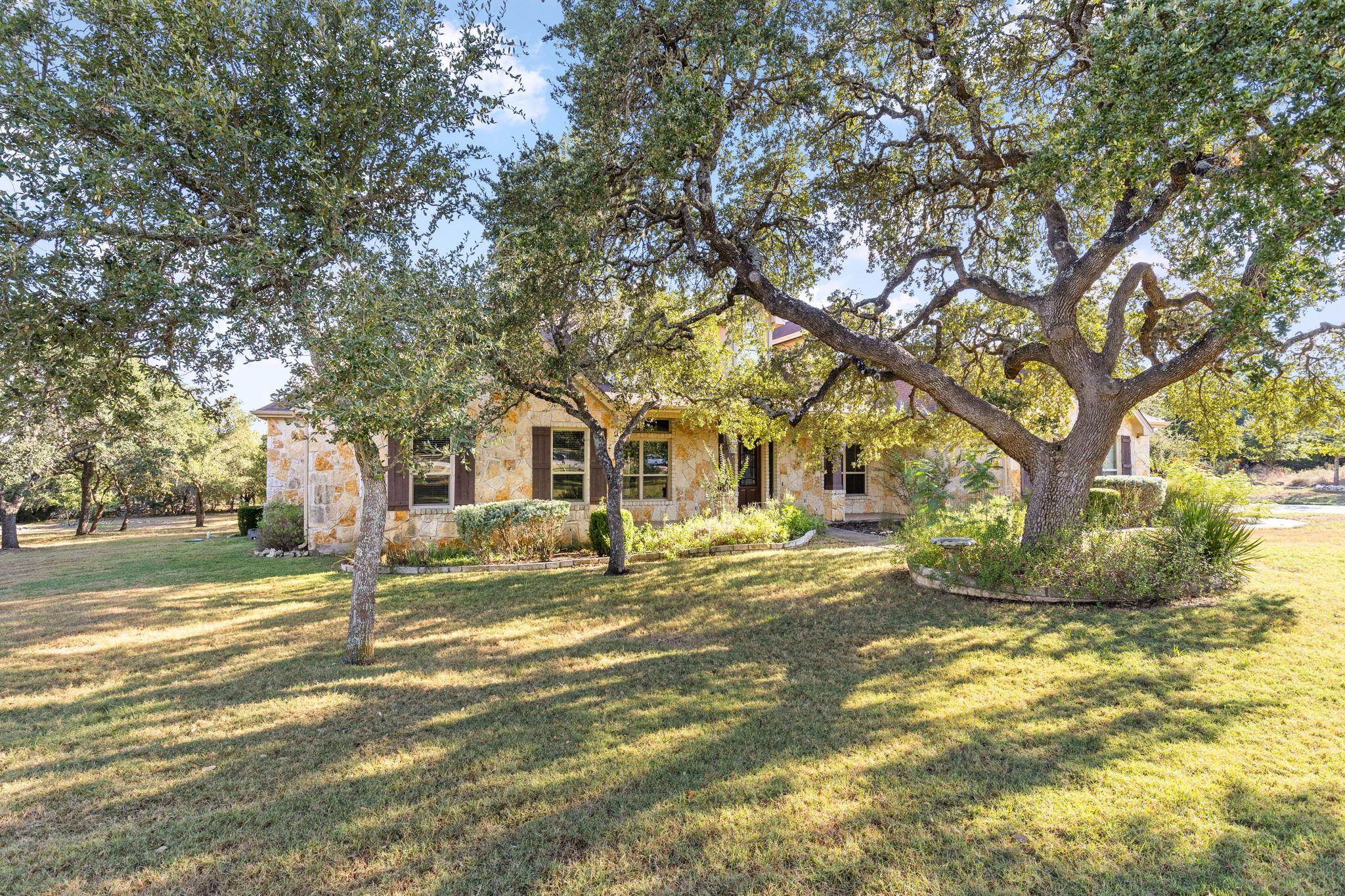 Stone house framed by mature oak trees, with a broad lawn and curved planting beds.