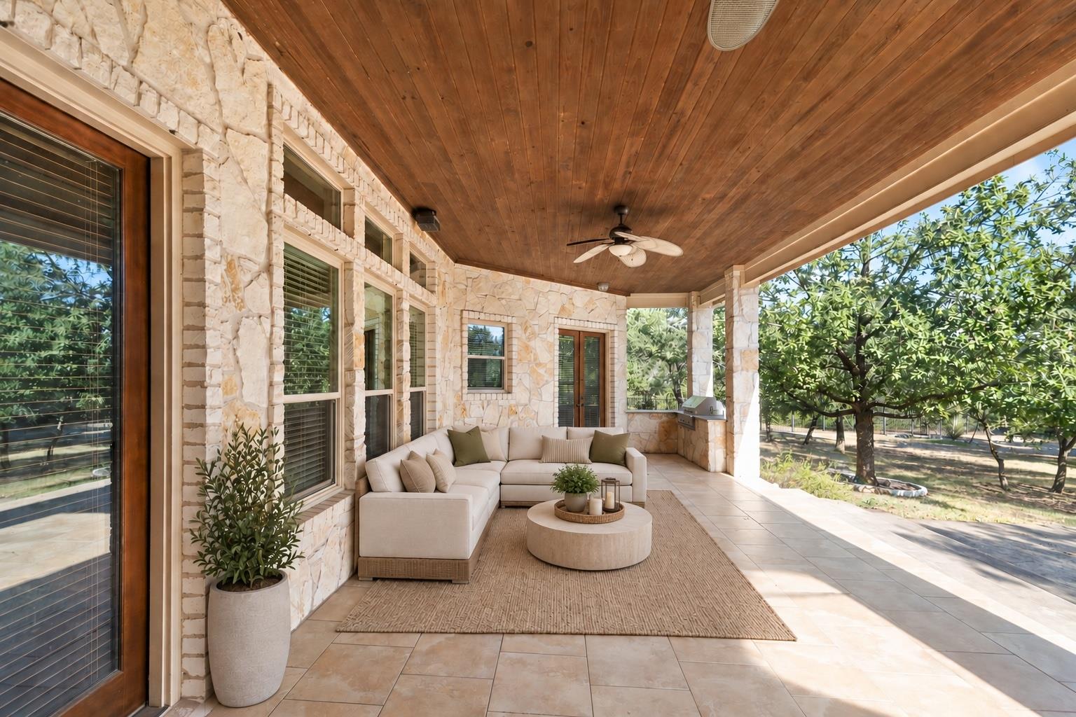 Covered stone patio with a wood ceiling, cream sectional, and views of a leafy yard.