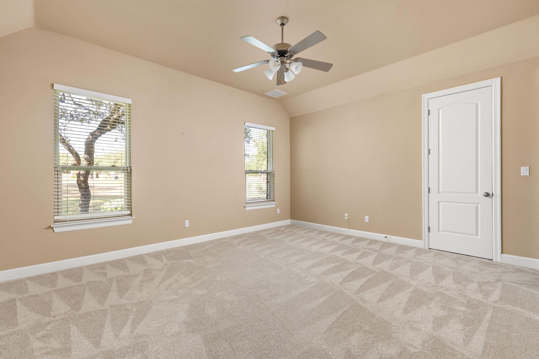 Empty carpeted room with tan walls, two windows, and a ceiling fan.