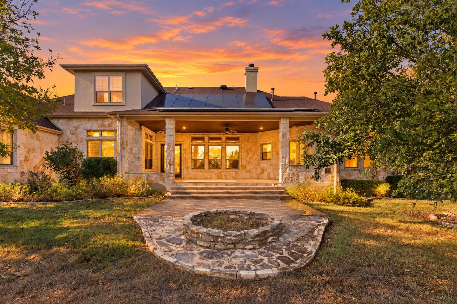 Stone house with a covered patio and solar panels behind a circular stone fire pit at dusk.