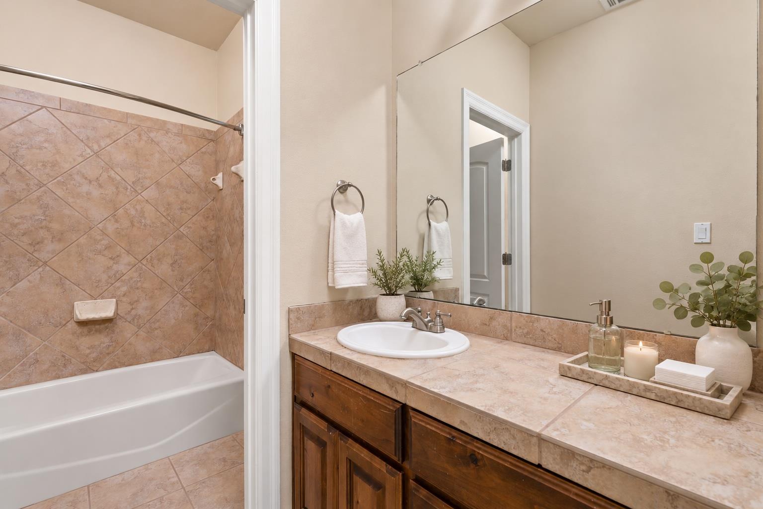 Bathroom with tan tiled tub surround and vanity, white sink, dark wood cabinets, and large mirror.