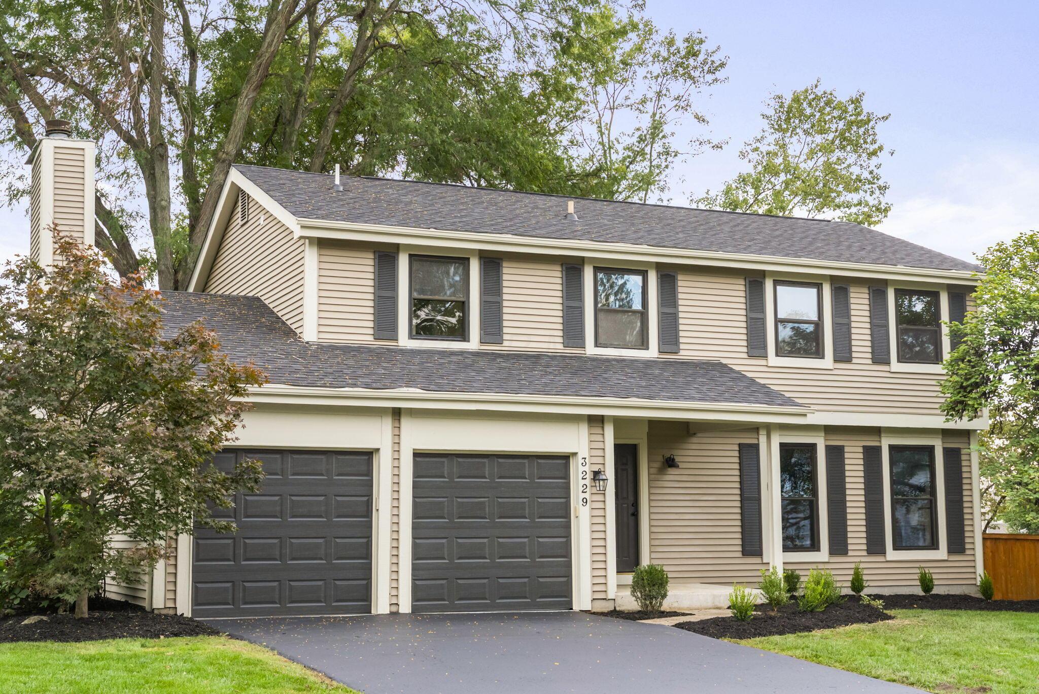 Two-story beige house with a double garage, dark shutters, and landscaped front yard.