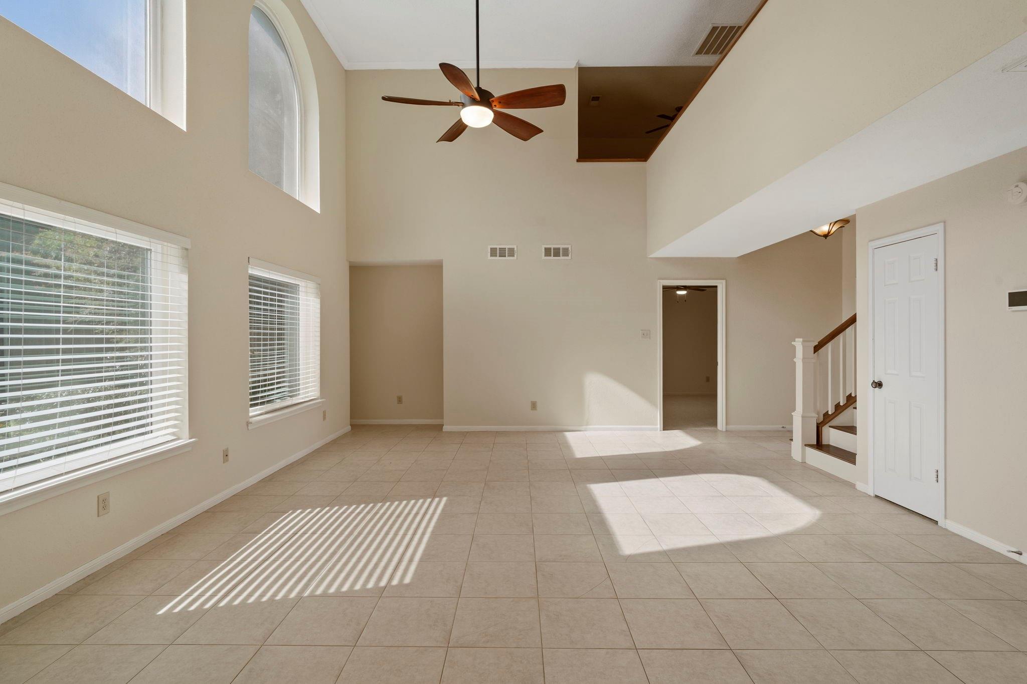 Empty double-height living room with tiled floors, tall windows, ceiling fan, and staircase.