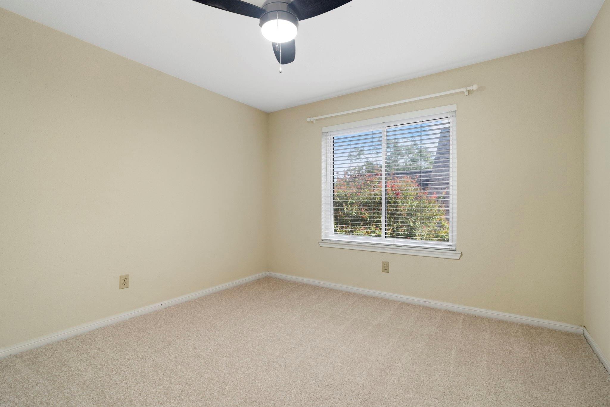 Empty carpeted bedroom with beige walls, a ceiling fan, and a window with white blinds.