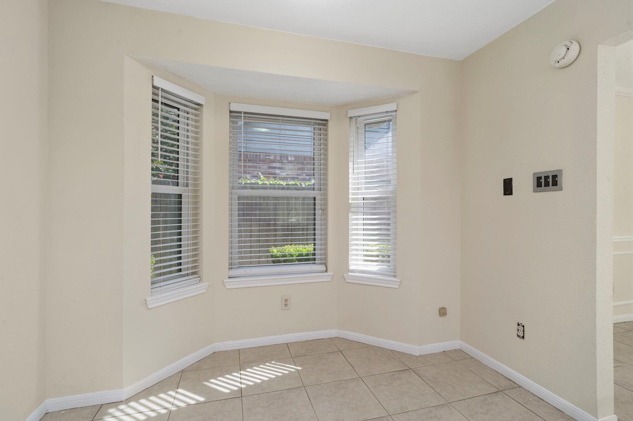Empty interior nook with three bay windows, white blinds, and sunlight across the tiled floor.