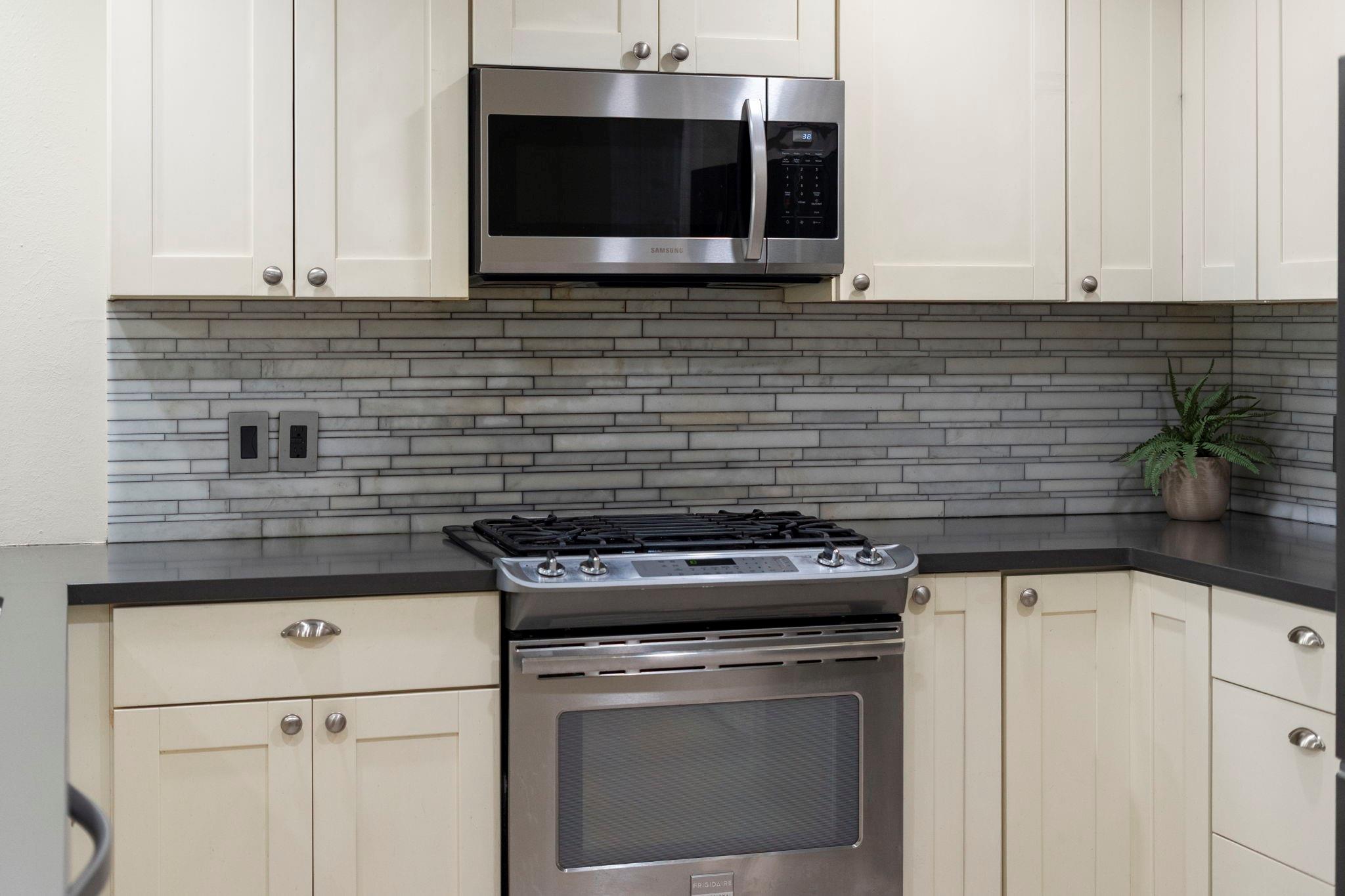 Kitchen with cream cabinets, gray tile backsplash, stainless gas range, and microwave.