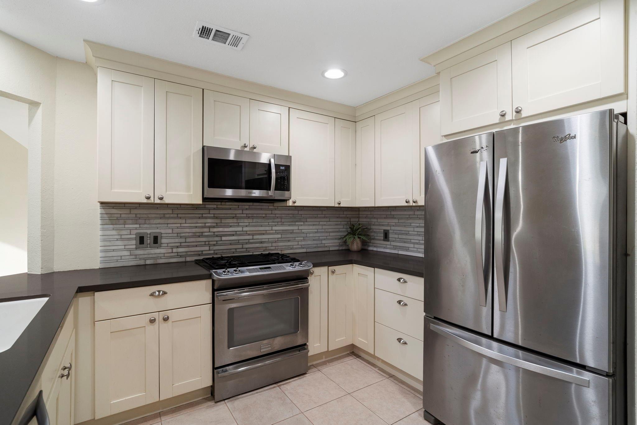 Kitchen with cream cabinets, dark counters, gray tile backsplash, and stainless steel appliances.