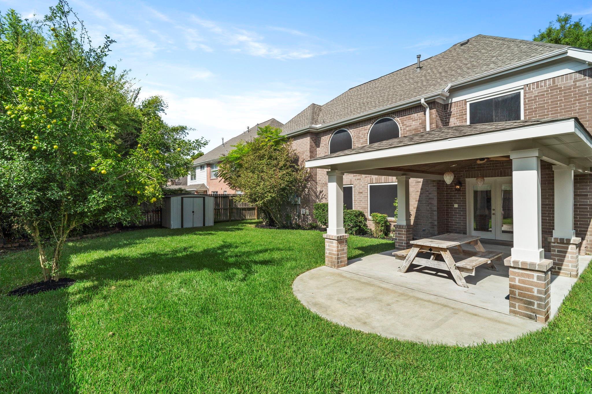 Brick home’s backyard with a covered patio, picnic table, lawn, fruit tree, and garden shed.