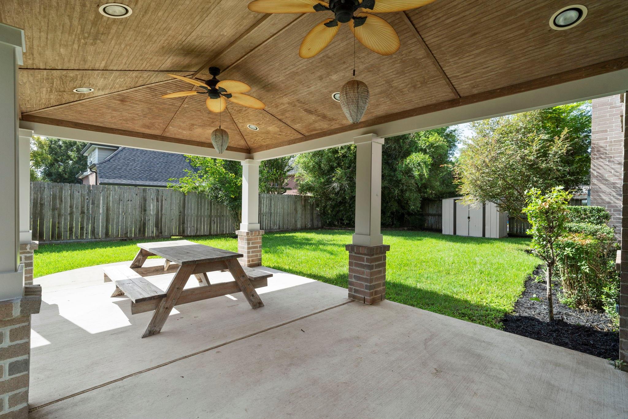 Covered backyard patio with a wood ceiling and ceiling fans, overlooking a fenced lawn.