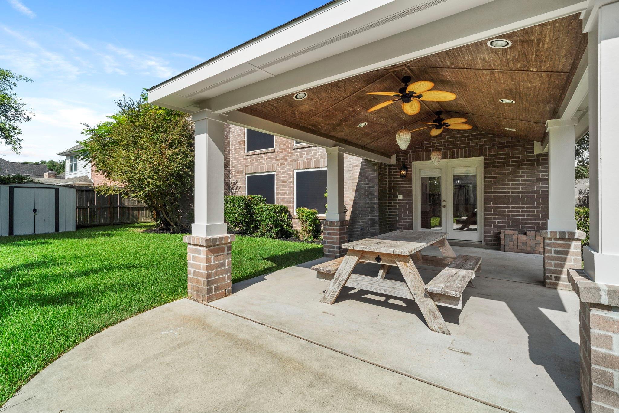 Covered brick patio with a wood-paneled ceiling, two ceiling fans, and a picnic table beside the lawn.